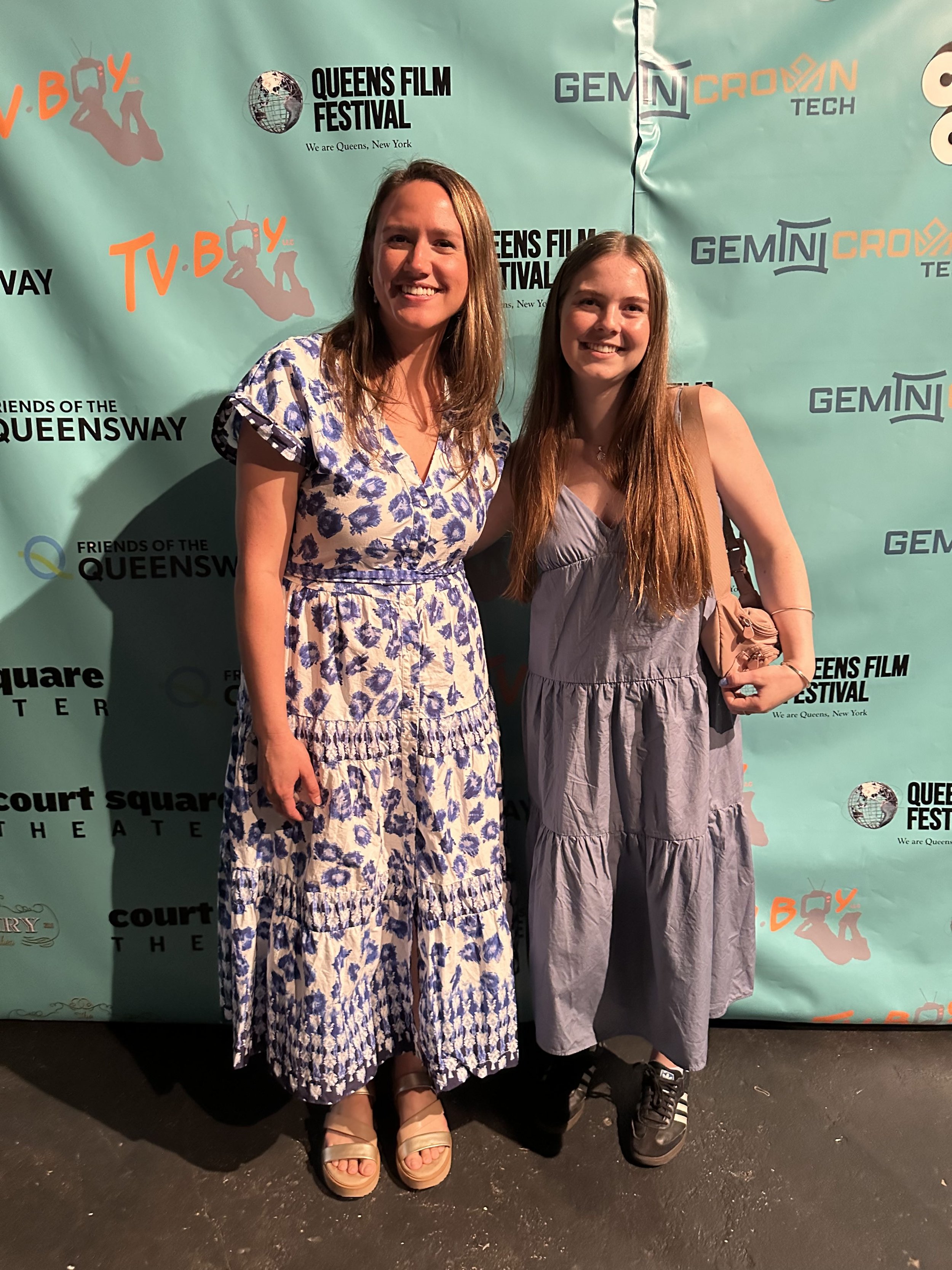 Two women standing together at the Queens Film Festival, posing in front of a teal backdrop with event logos.