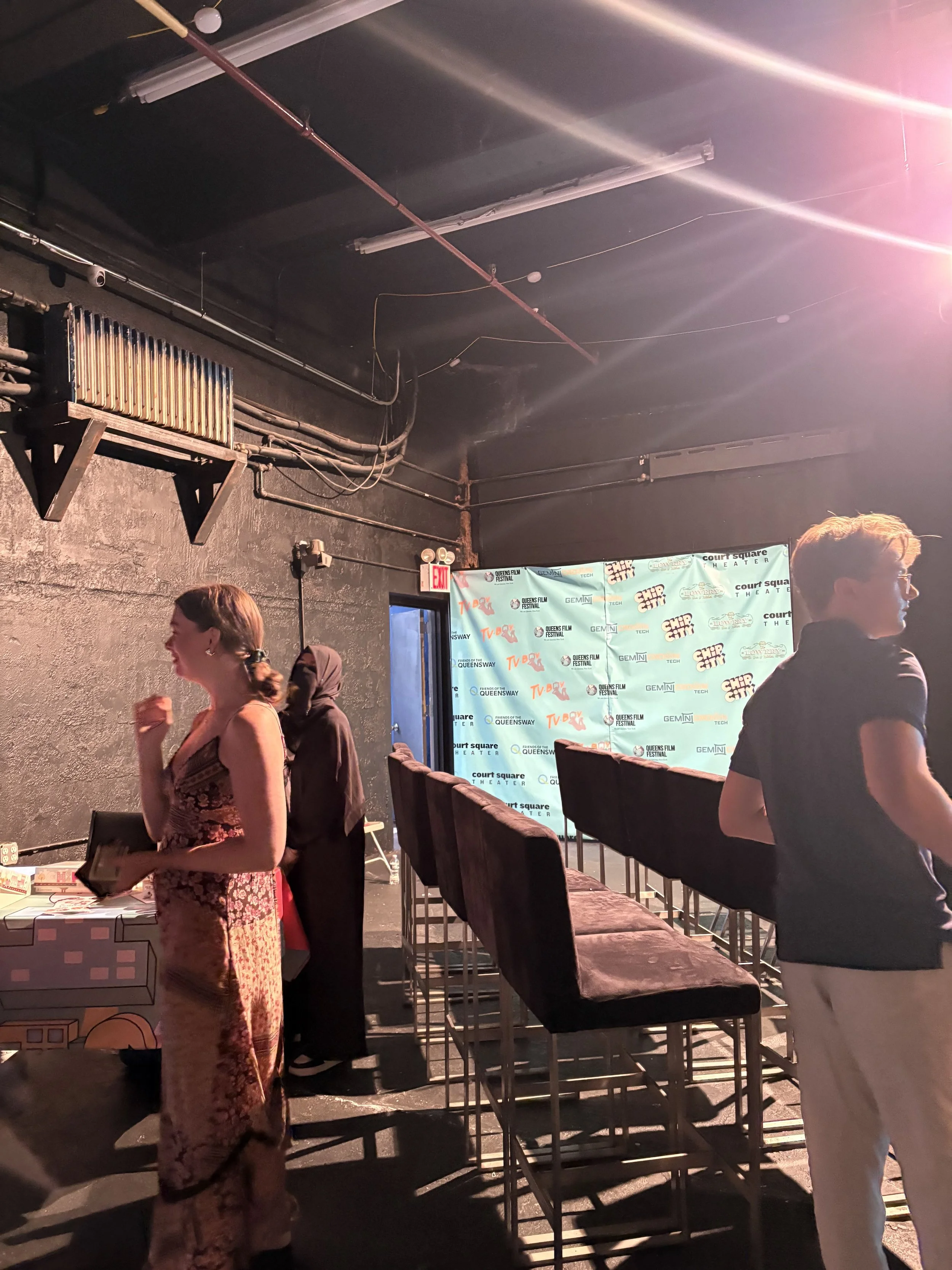 An indoor scene at a venue with a black wall, a screen displaying logos, and several people standing near rows of chairs. The ceiling has exposed pipes and lighting.
