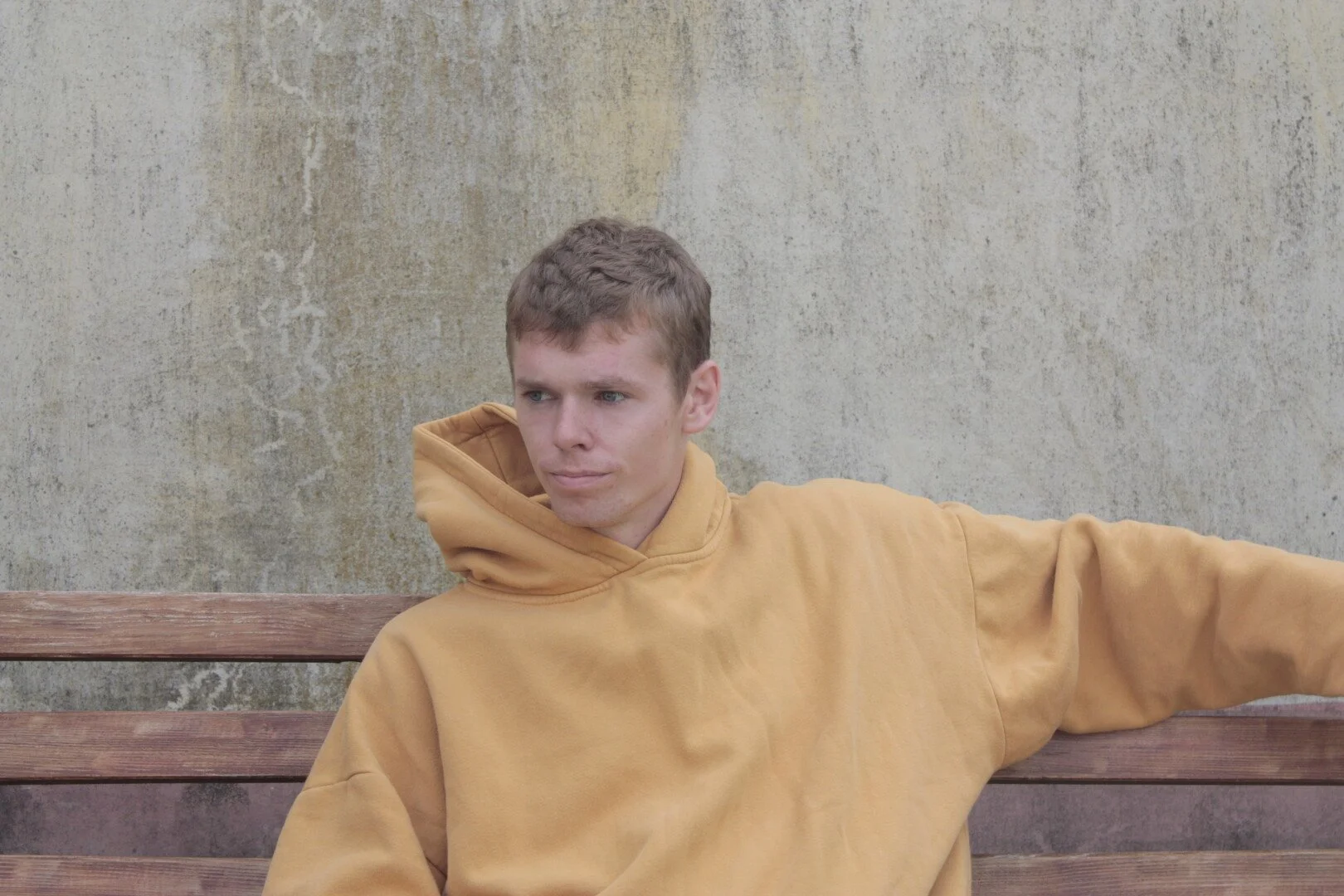 A young man in a yellow hoodie sitting on a wooden bench against a weathered concrete wall.
