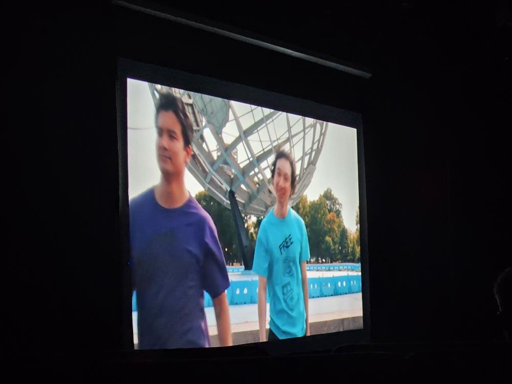 Projection screen displaying a photo of two young people outdoors near a large spherical metallic structure, with trees and a pool in the background.