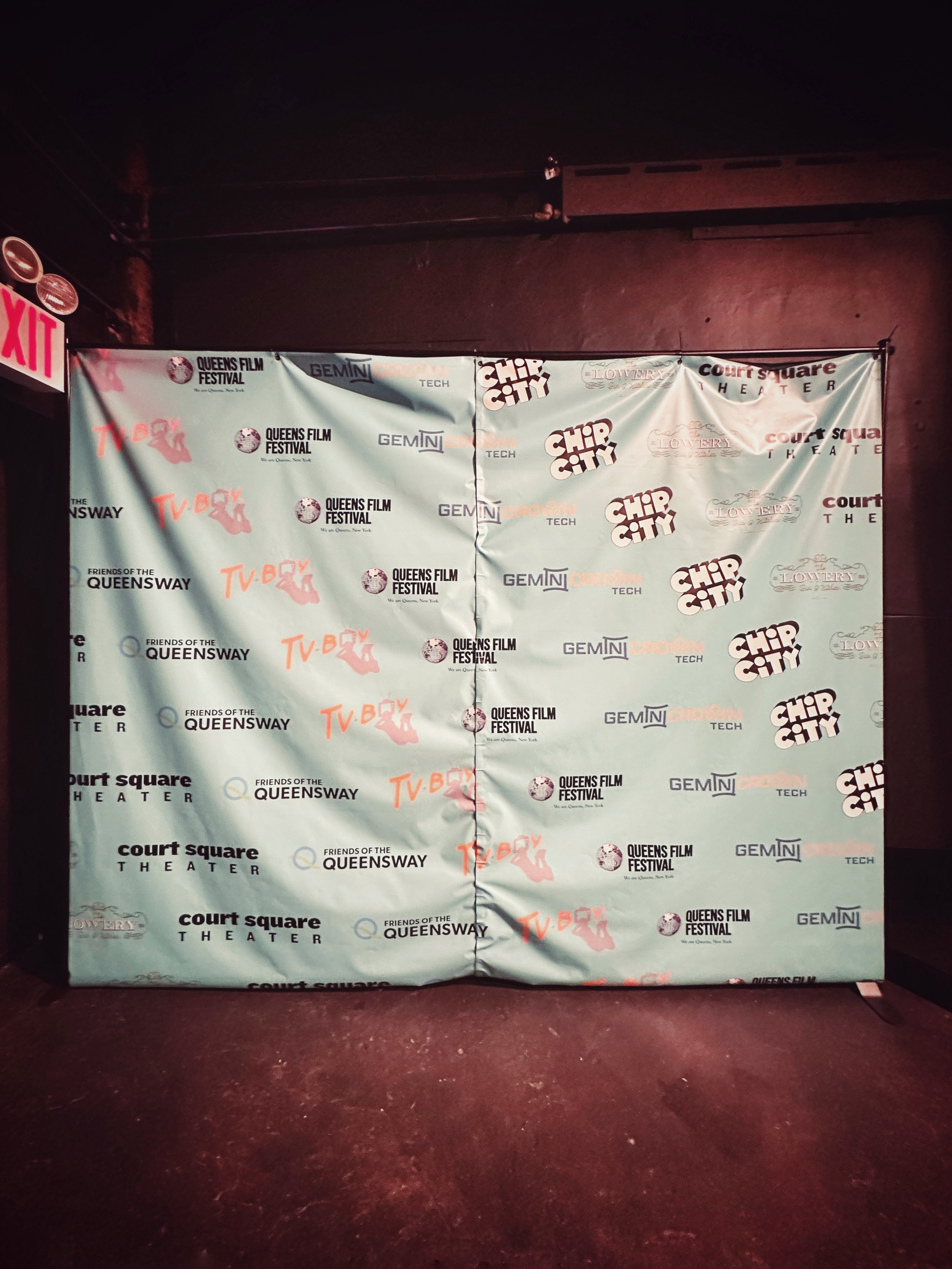 Step and repeat banner with logos for Queens Film Festival, Chip City, Ghosts of the Lower, Queensway, and Court Square Theater, set against a dark wall with red lighting.