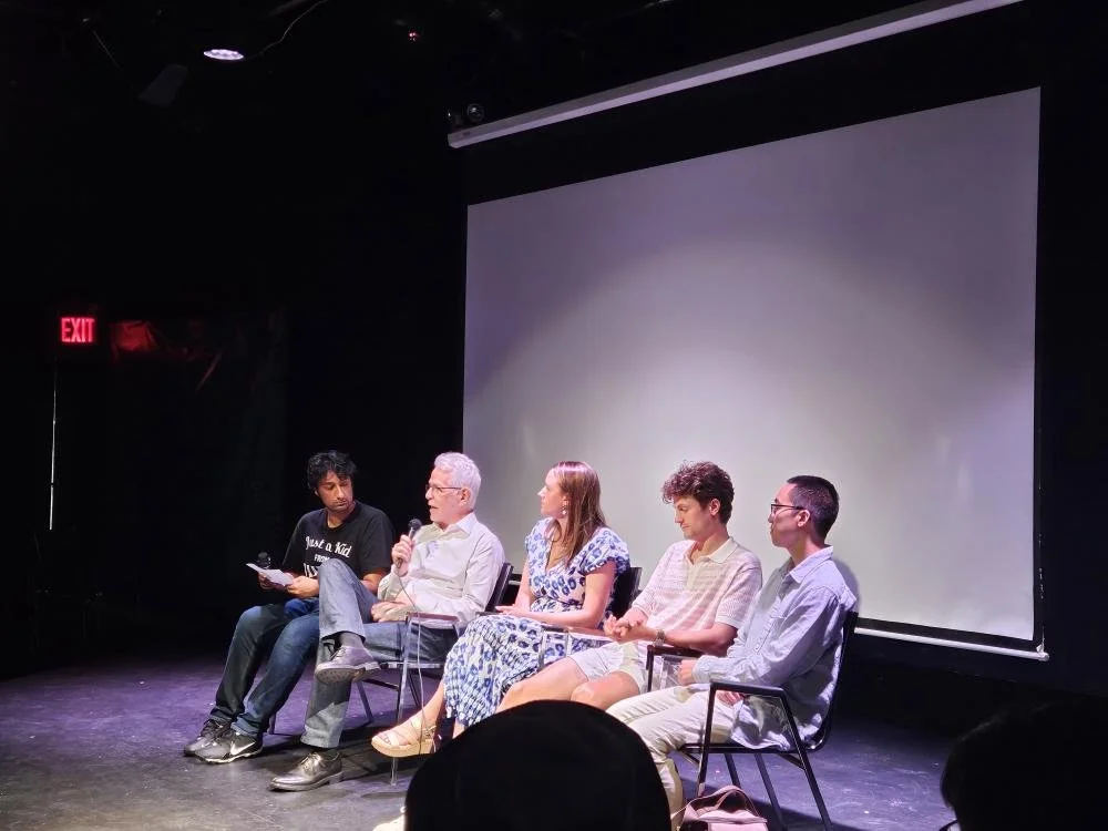 Five people seated on a stage in front of a large blank screen, participating in a panel discussion or presentation. One person is speaking into a microphone, while the others listen.