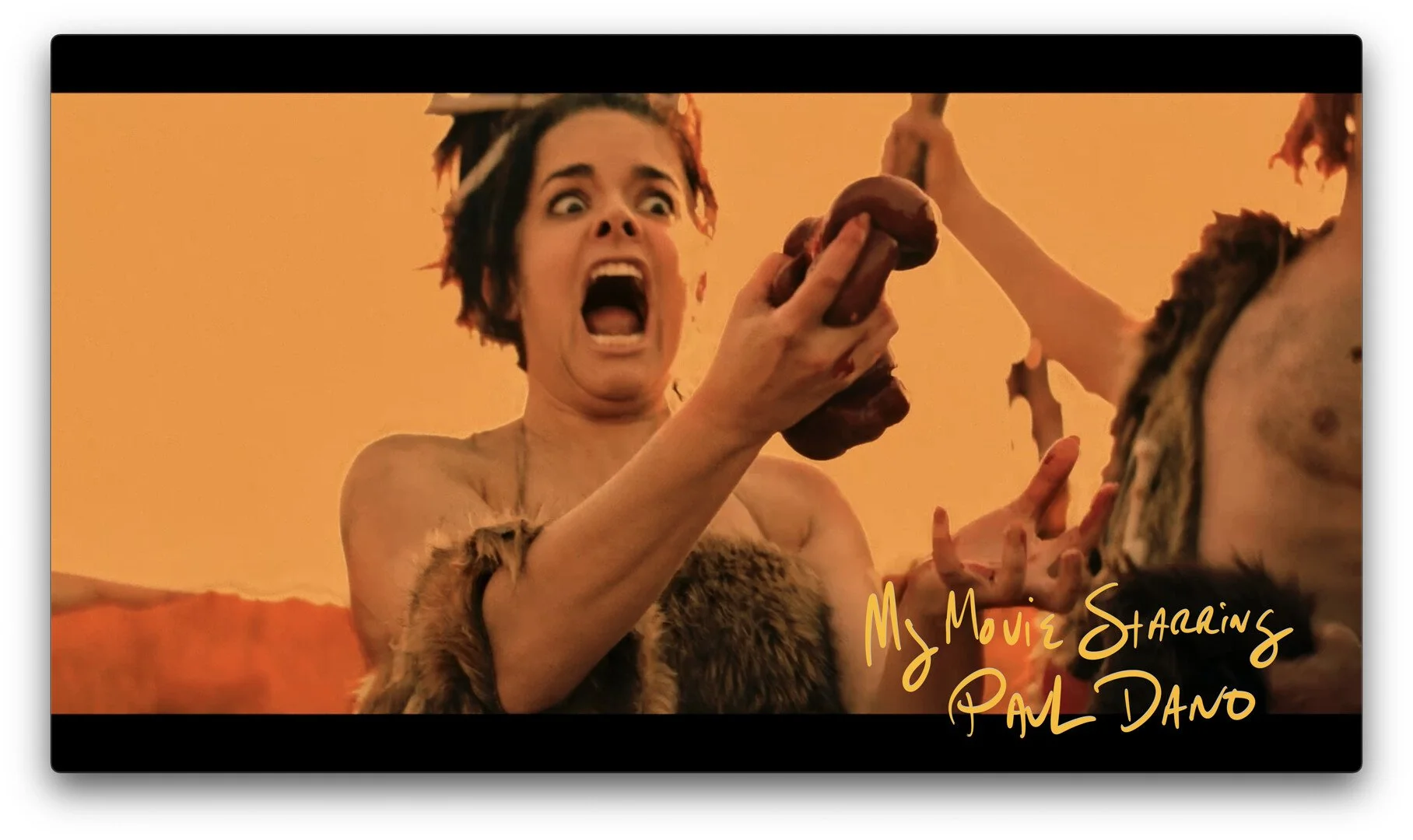 A woman with short dark hair and a surprised expression, holding a large object that appears to be a cluster of primitive-looking objects, wearing fur and standing against an orange desert background, with text reading 'My Movie Starring Paul Dano'.