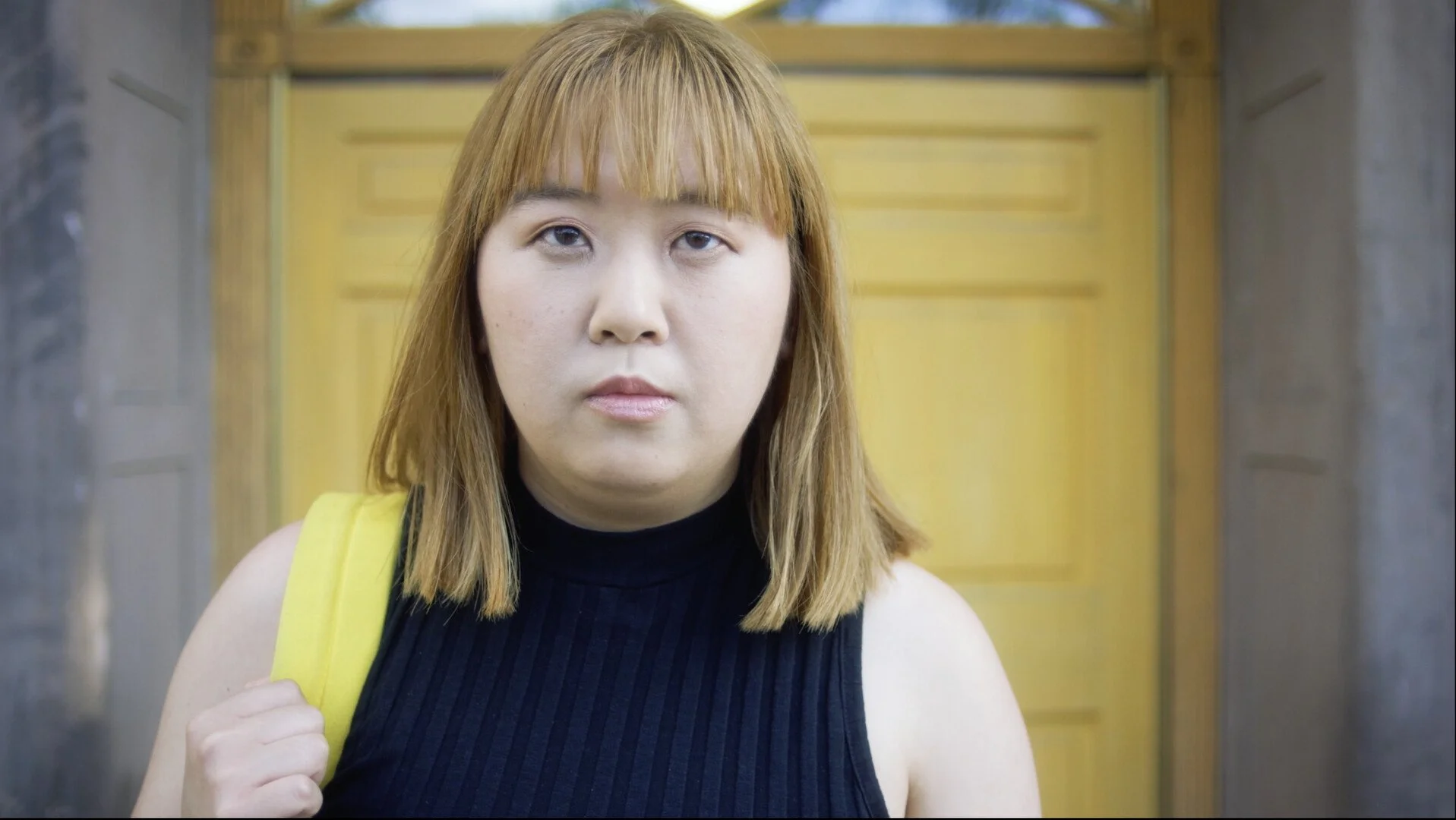 Woman with shoulder-length blond hair wearing a black sleeveless top and yellow backpack standing in front of a yellow door.