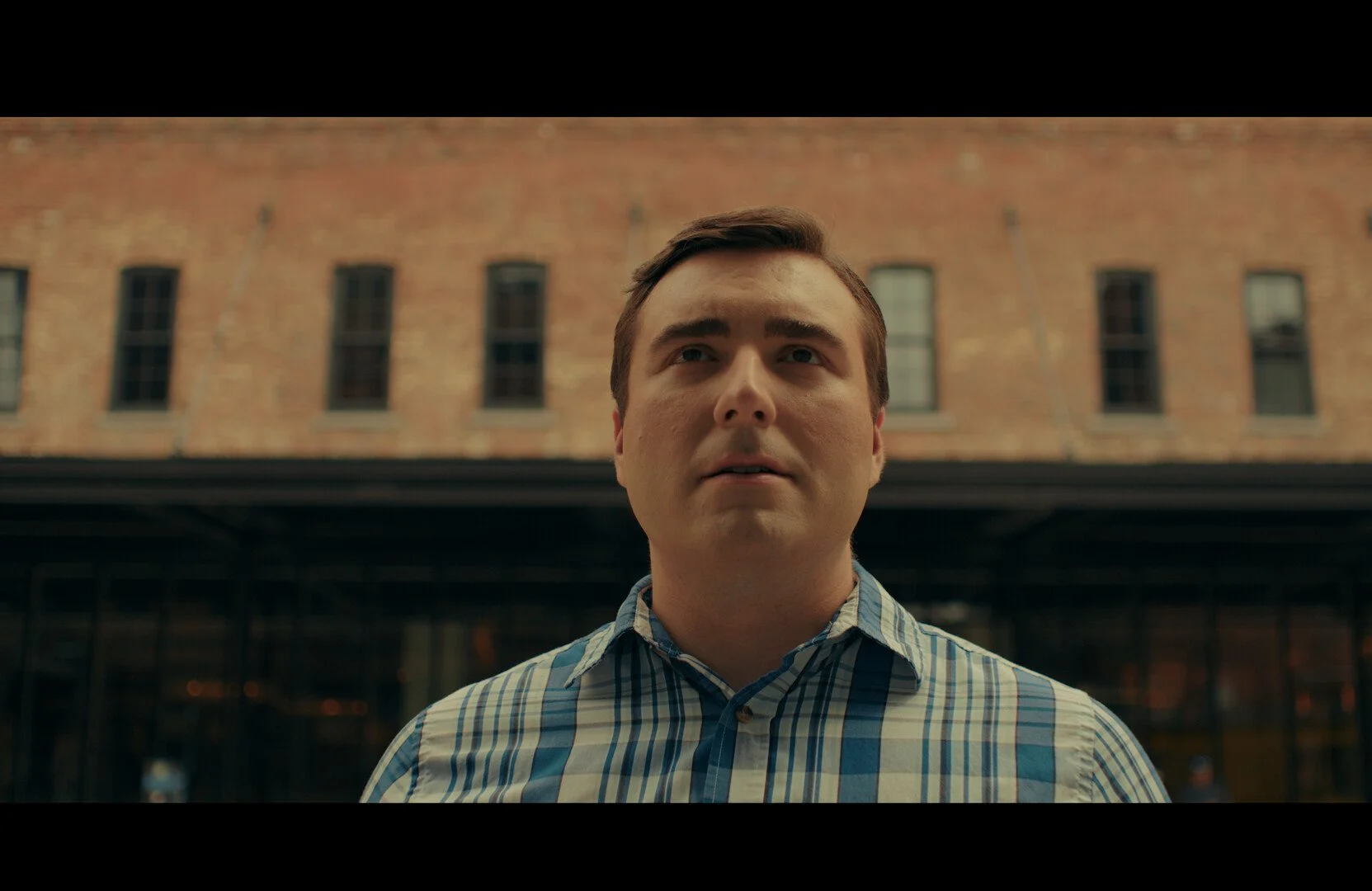 A man with short dark hair and wearing a checkered shirt standing outdoors in front of a brick building with multiple windows.