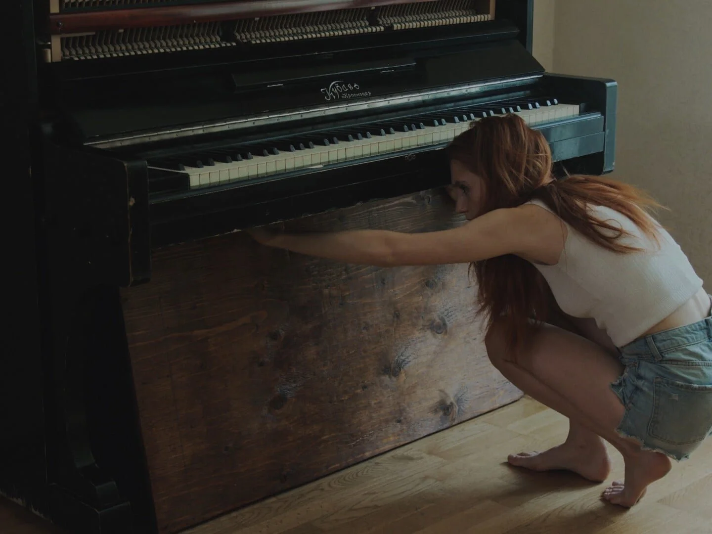 A woman with long red hair crouching barefoot next to a black upright piano with a wooden panel, stretching her arm to touch the keys.