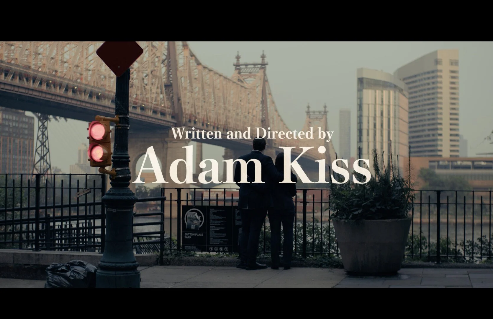 Title screen of a film showing two men in suits standing by a fence near a river, with the Brooklyn Bridge and city skyline in the background, and text indicating it was written and directed by Adam Kiss.