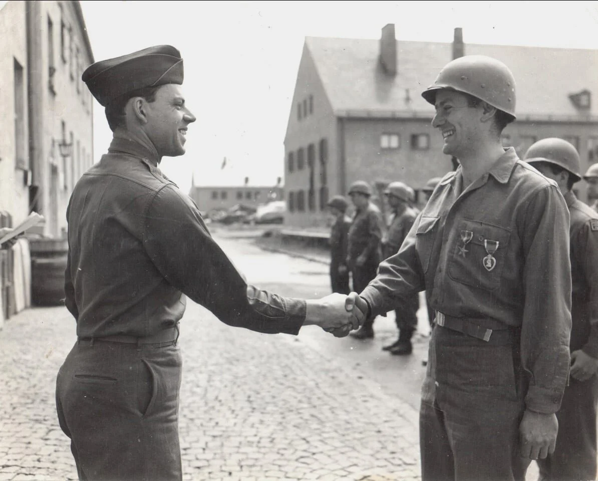 Two soldiers, one in a military uniform with a cap and the other in a uniform with a helmet, shake hands and smile, with several soldiers in formation in the background.