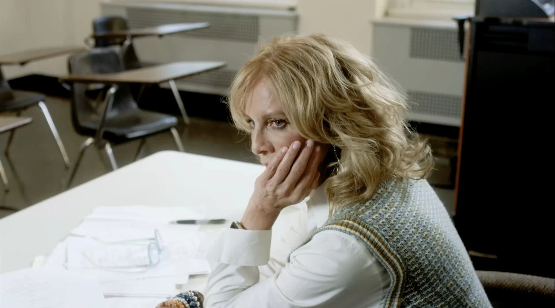A woman with blonde curly hair sitting at a desk in a classroom or office, resting her face on her hand, with papers and a pen in front of her.