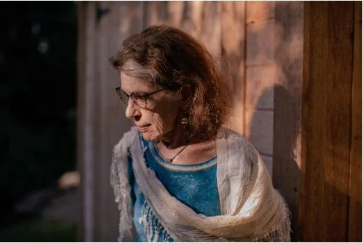 A woman with glasses and brown hair leaning against a wooden wall, looking downward.