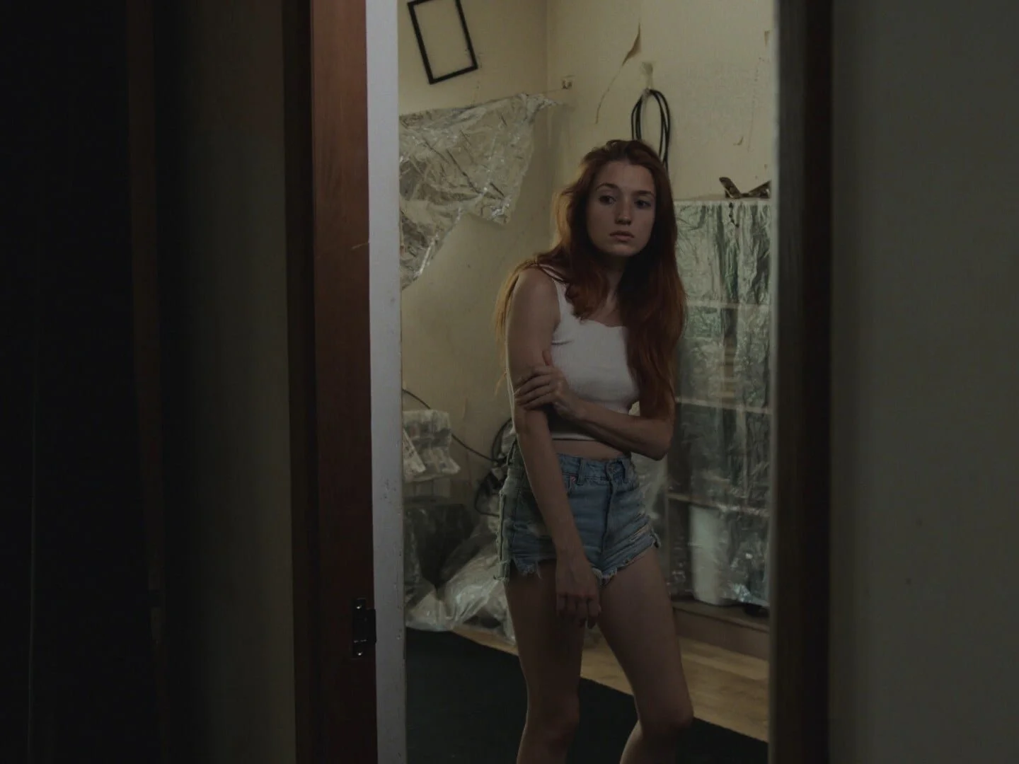 A young woman with long red hair, wearing a white crop top and denim shorts, stands in a room reflected in a mirror, with a concerned or pensive expression on her face.