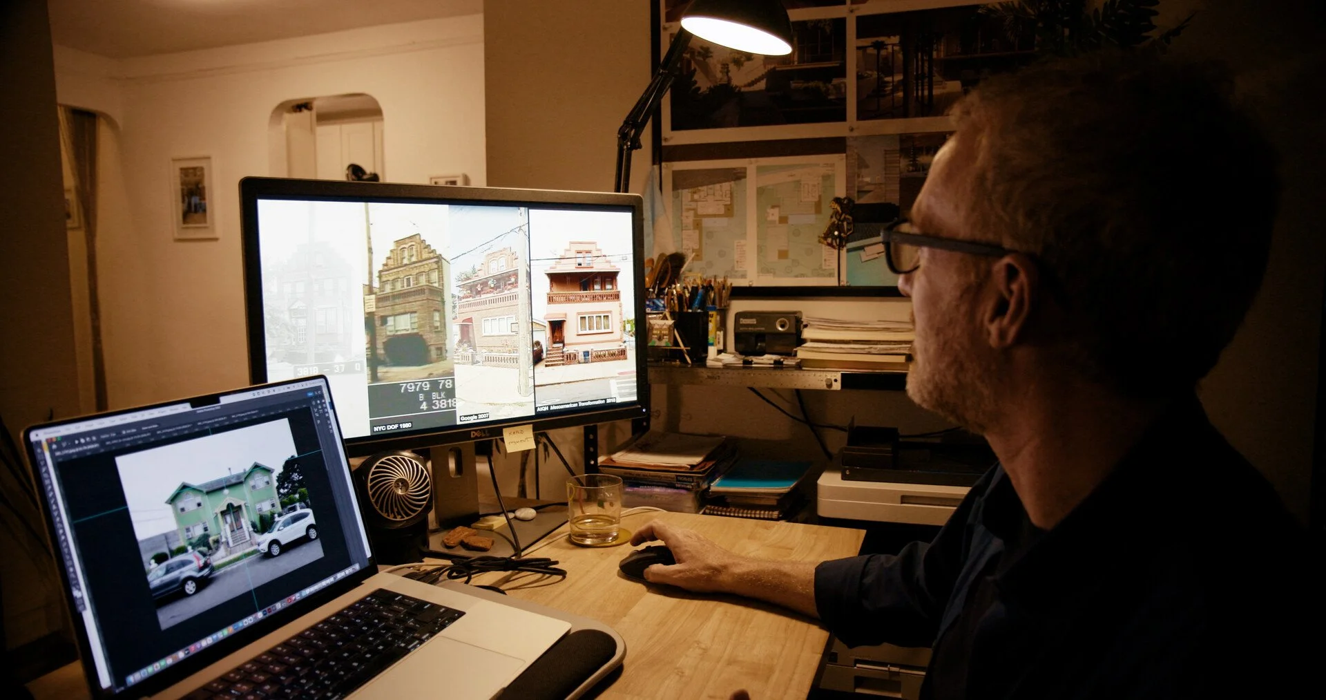 A man working at a desk with a laptop and monitor, analyzing images of houses for security or design purposes, in a dimly lit room.