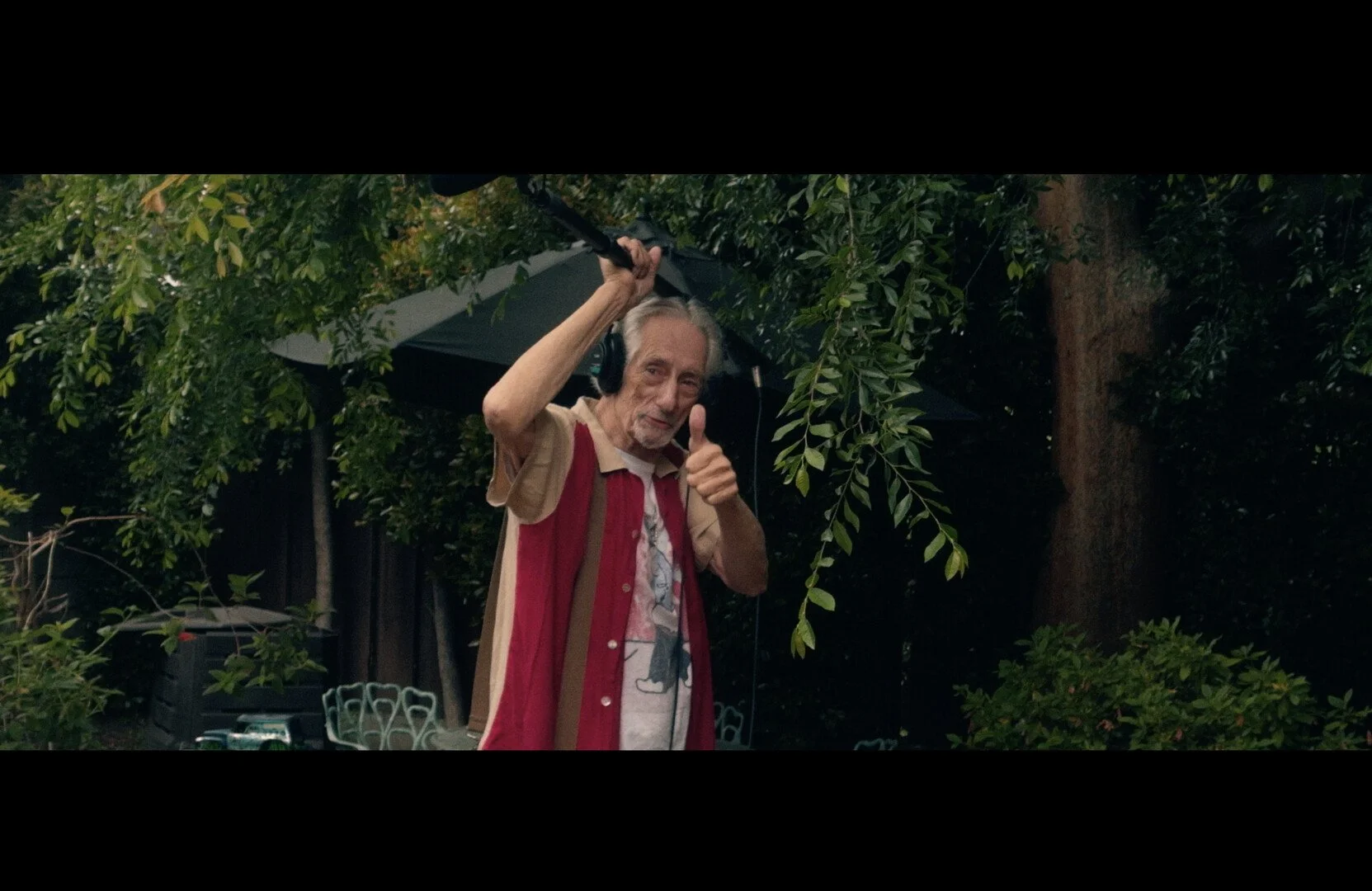 An elderly man wearing headphones and a colorful shirt giving a thumbs-up while holding an umbrella in a lush, green outdoor setting.