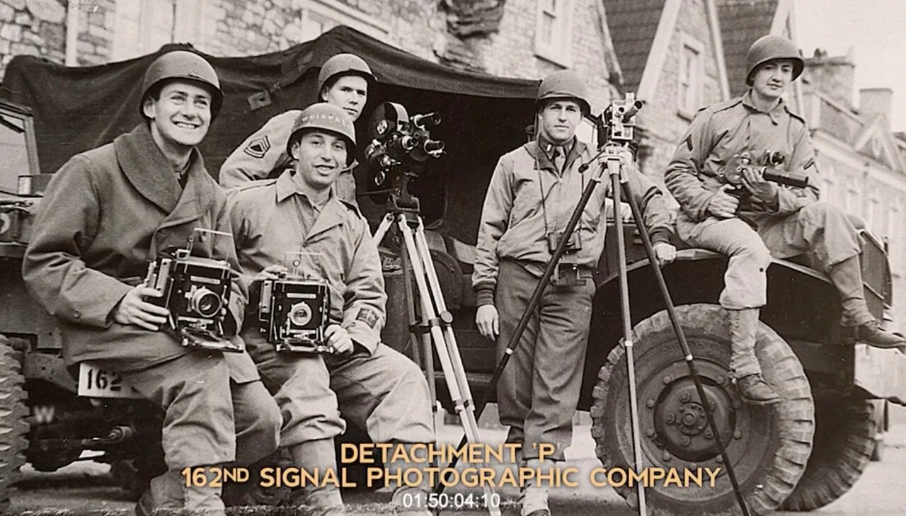 Black and white photo of five soldiers in military uniforms with helmets, holding cameras and photography equipment, posing in front of a military truck with buildings in the background.