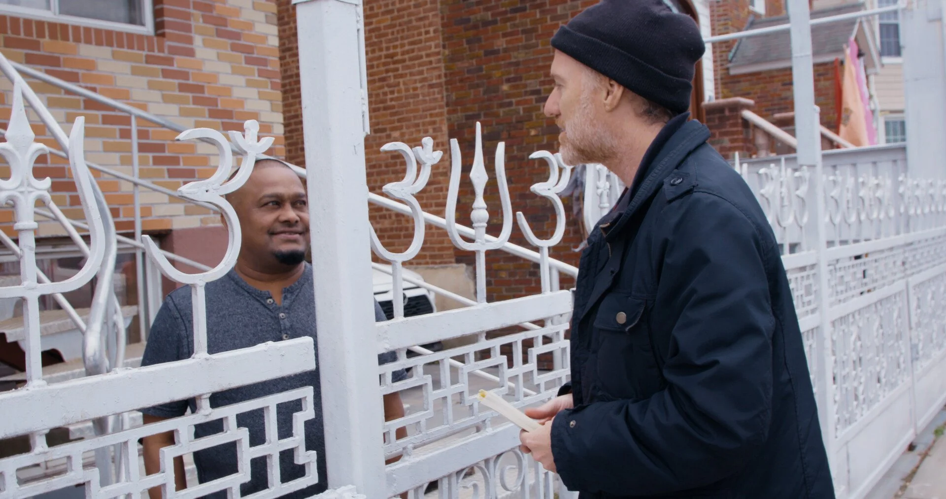 Two men having a conversation at a porch gate. One man stands outside the gate holding a paper, and the other man stands behind the gate in front of a brick house.