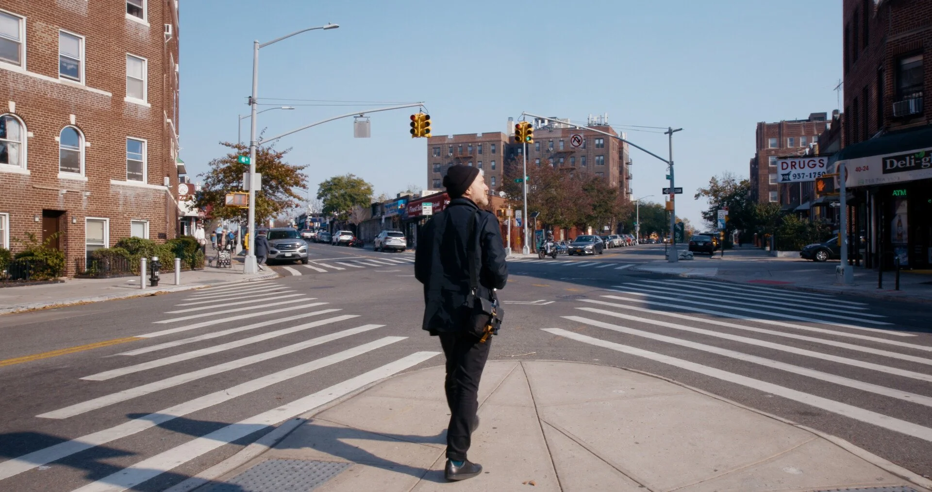 A man crossing a city street at a crosswalk with buildings and shops on either side, traffic lights, and cars in the background.