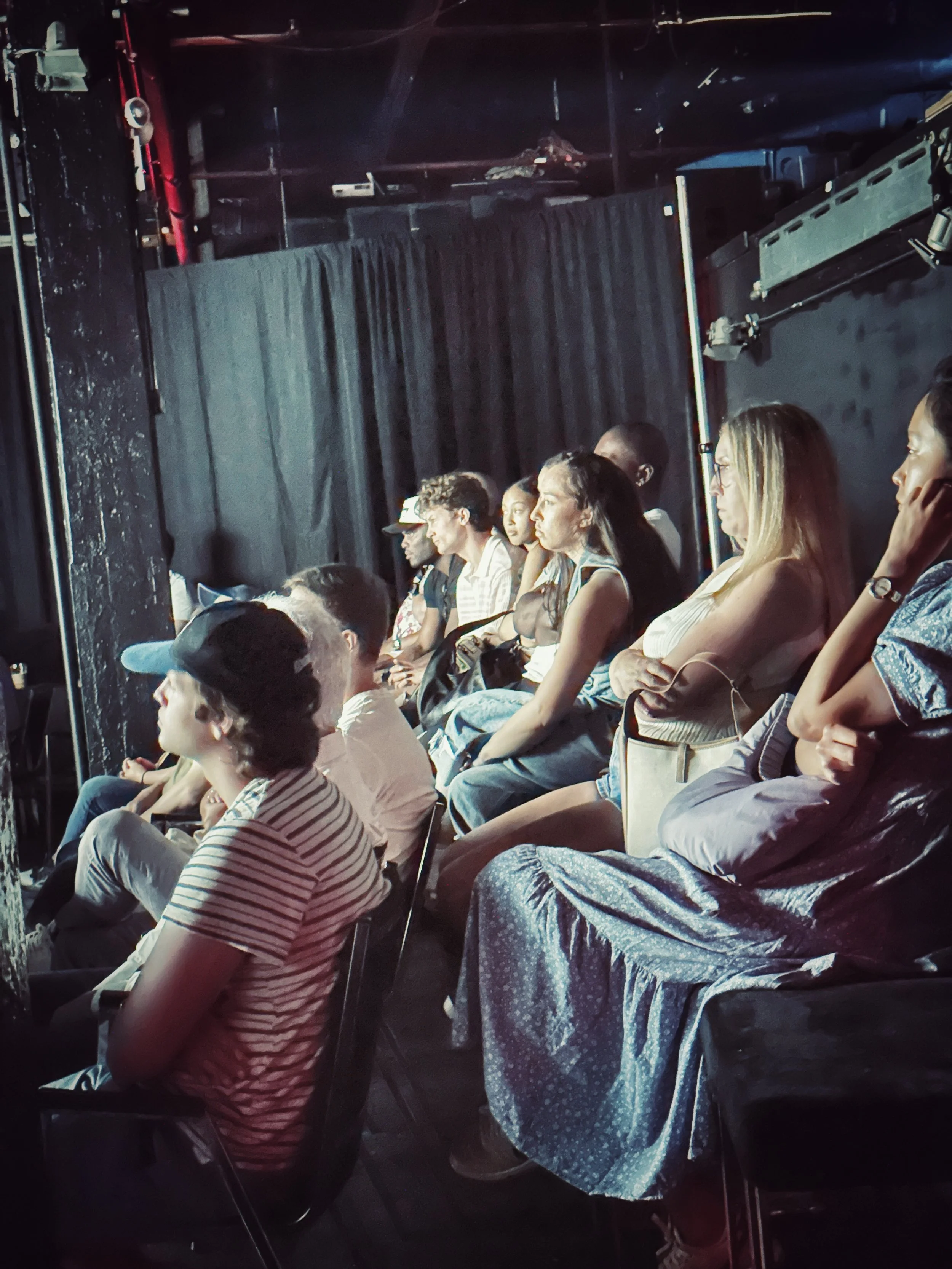 A group of people sitting and watching something in an indoor black box theater or performance space, with black curtains and stage equipment visible in the background.