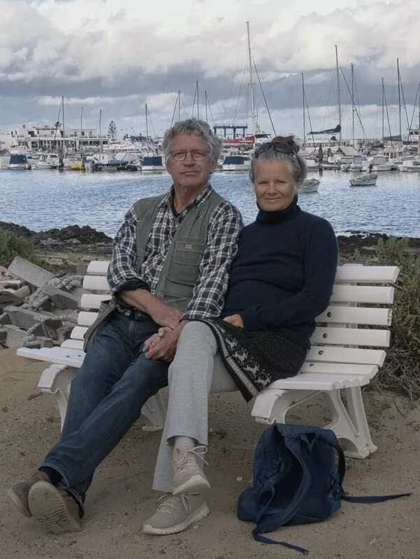 Older man and woman sitting close together on a white bench by a marina with boats and yachts in the background.