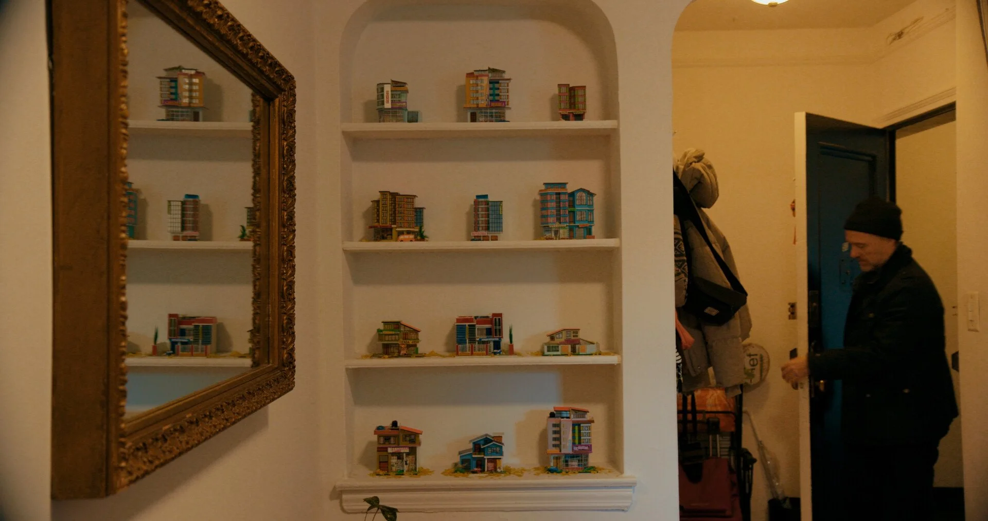 A woman in a black jacket and black beanie looking at miniature models of houses displayed on white built-in shelves next to a large ornate mirror in a room with beige walls.