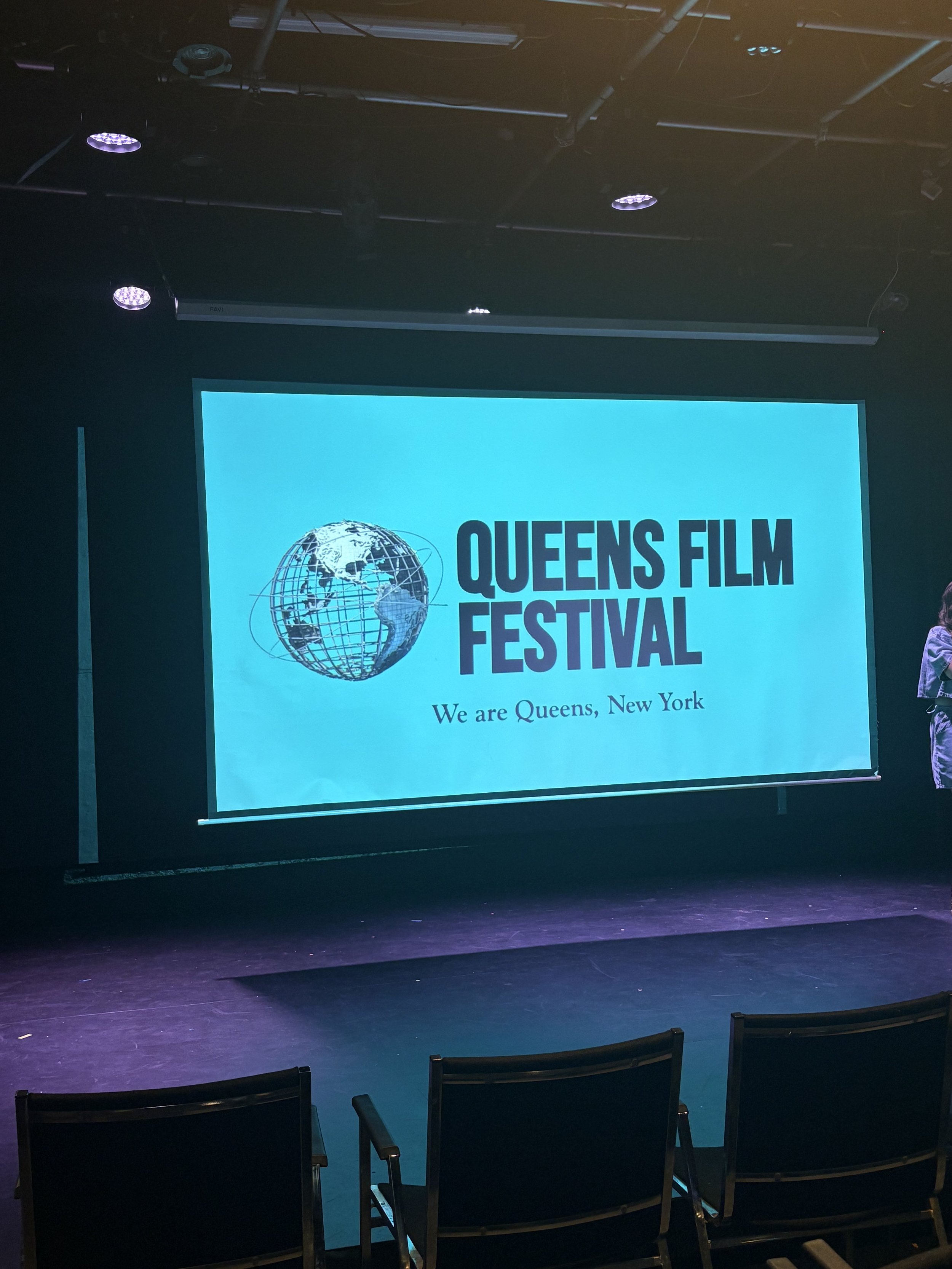 A large screen on a stage displaying the logo for Queens Film Festival, which features a globe with a world map and the text 'QUEENS FILM FESTIVAL' and 'We are Queens, New York'.