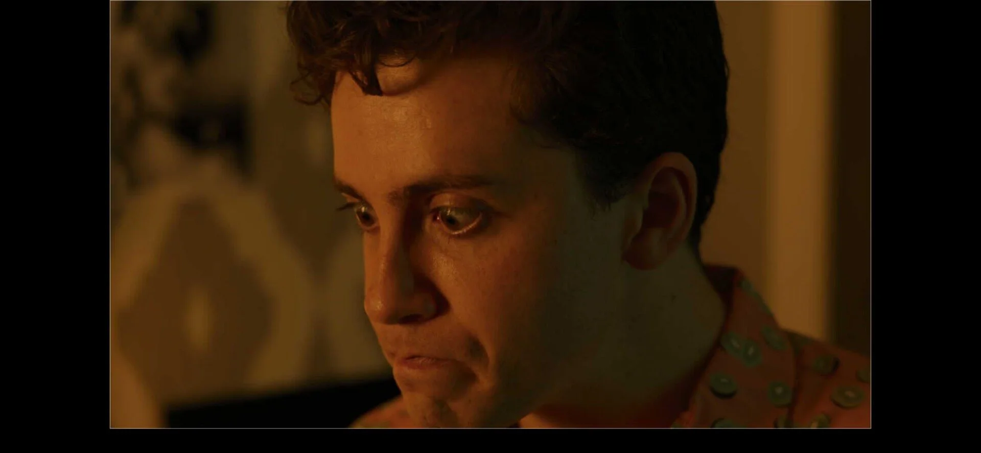 A close-up of a young man with curly dark hair, pale skin, and green eyes, wearing a patterned shirt with circular designs, looking downward in a dimly lit room.