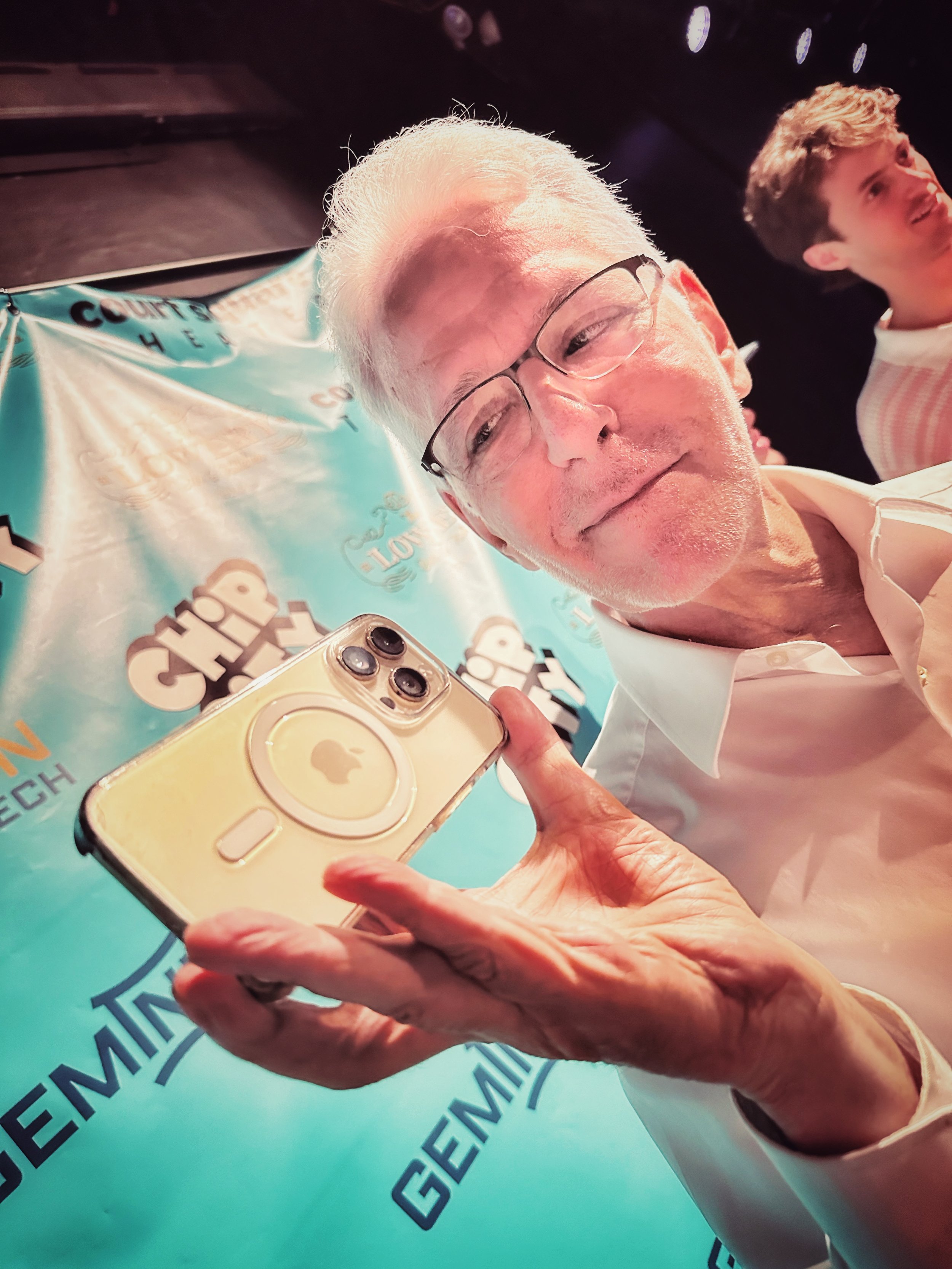 An elderly man with glasses holding a yellow iPhone with a pop socket, smiling at the camera. In the background, a younger man is smiling, and a blue banner with text and logos is partially visible.