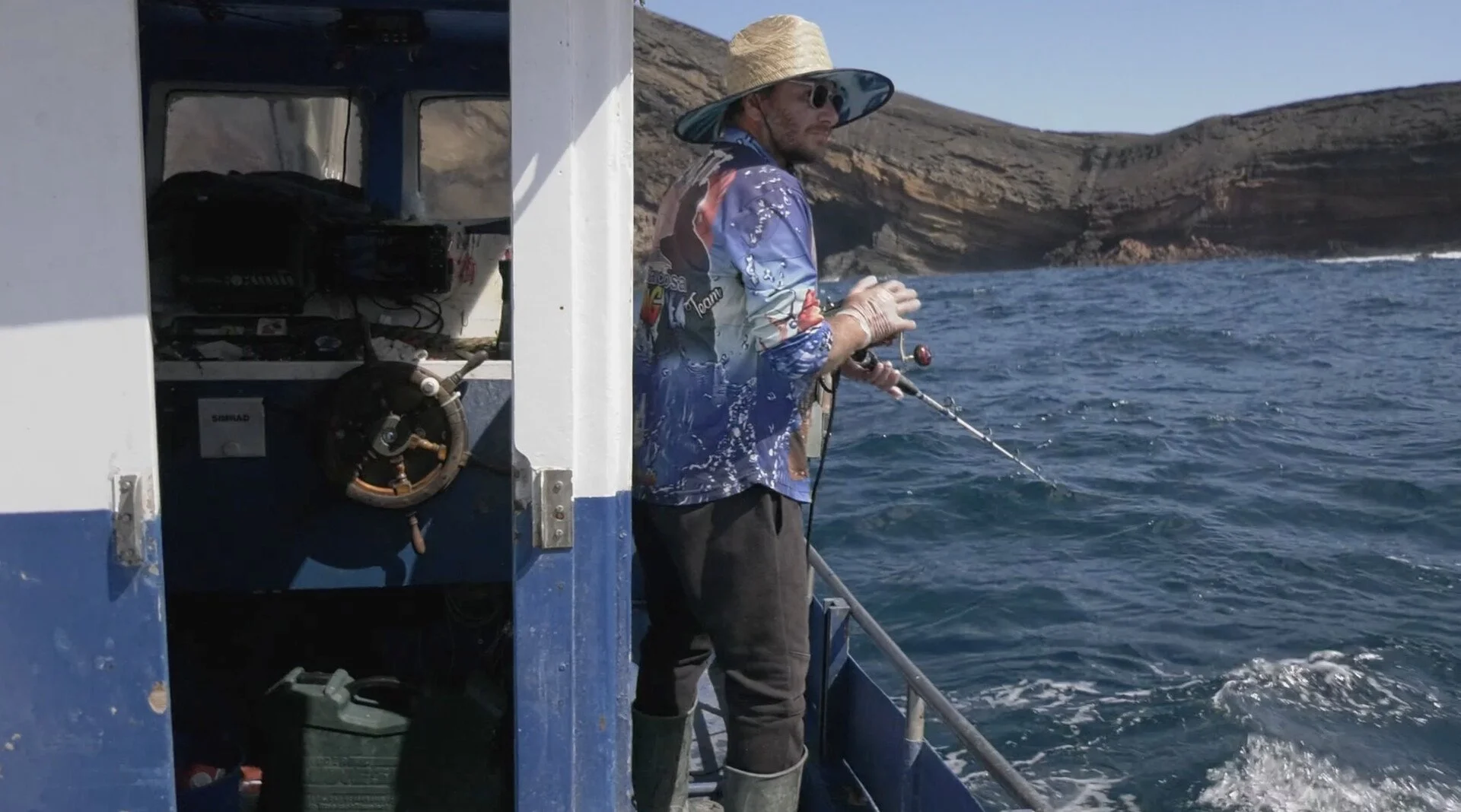 Man fishing on a boat near rocky coastline, wearing a large straw hat, sunglasses, a colorful fishing shirt, gloves, and rubber boots, holding a fishing rod over the water.