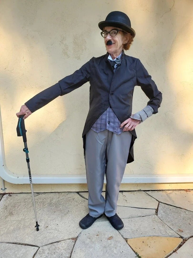 Person dressed as Charlie Chaplin with a cane, bowler hat, fake mustache, and glasses standing outside against a beige wall.
