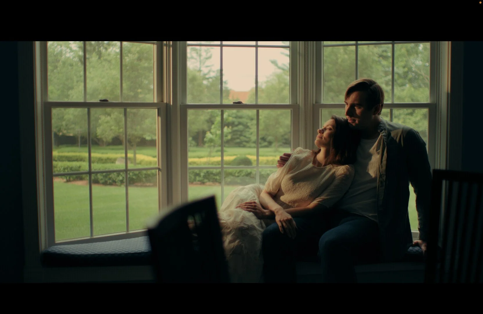 A couple sitting on a window seat by large windows, with a green garden outside, sharing an intimate moment with the woman resting her head on the man's shoulder.