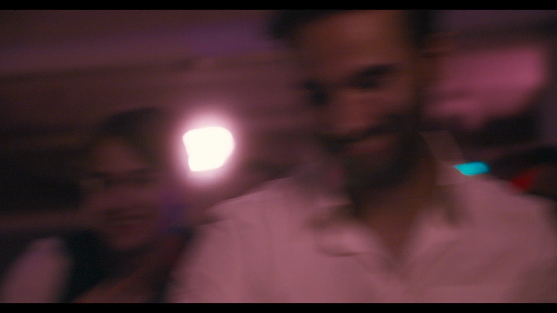 Two women in a dimly lit room with colorful lights, one woman with dark hair and a beard, wearing a white shirt, and the other woman in the background smiling with long dark hair.