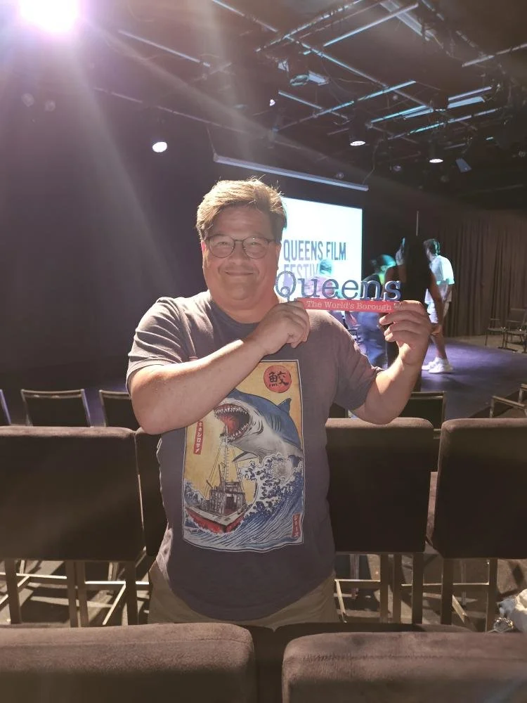 A man with glasses and a graphic T-shirt smiling and holding a sign that says "Queens, the World's Borough," at a film festival event in a theater setting.