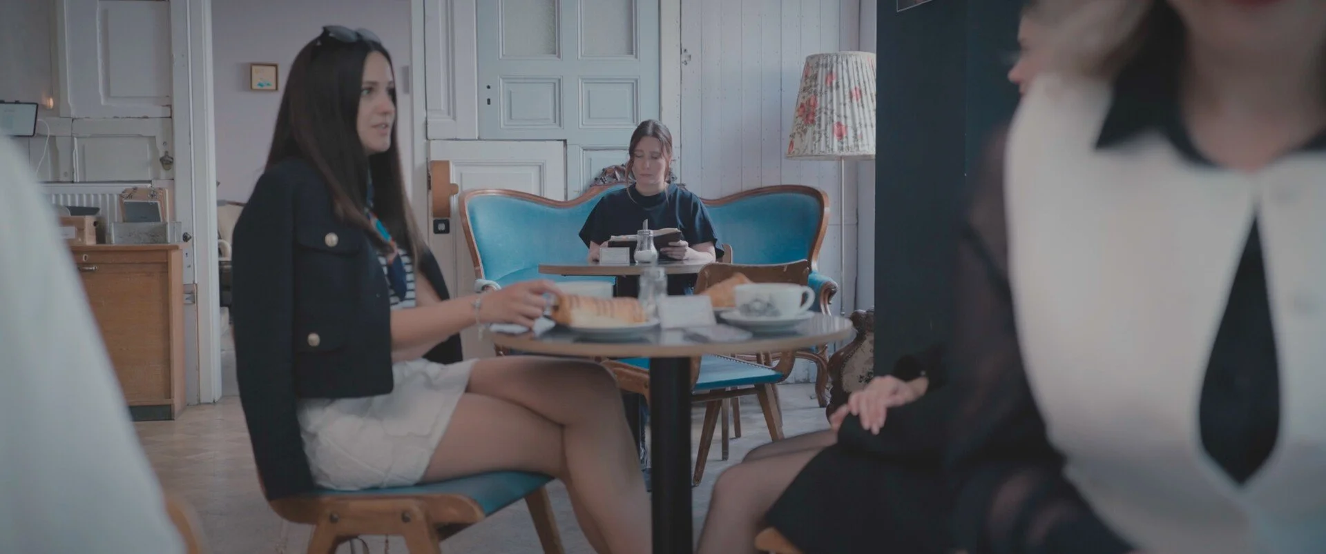 Three women sitting at a table in a cafe, two conversing with each other, the third woman reading a menu in the background.