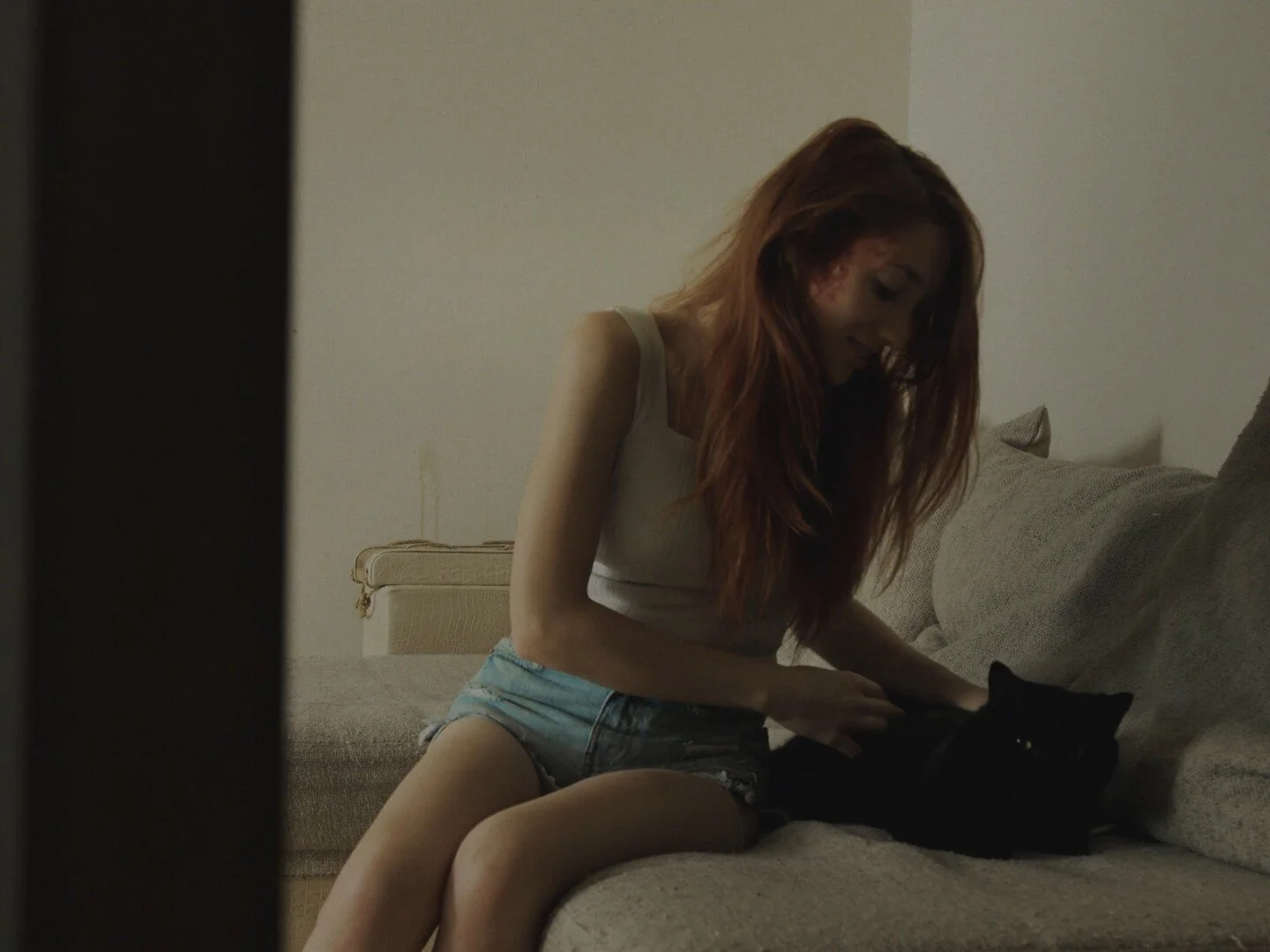 A woman with long red hair sitting on a beige couch, petting a black cat, in a dimly lit room.