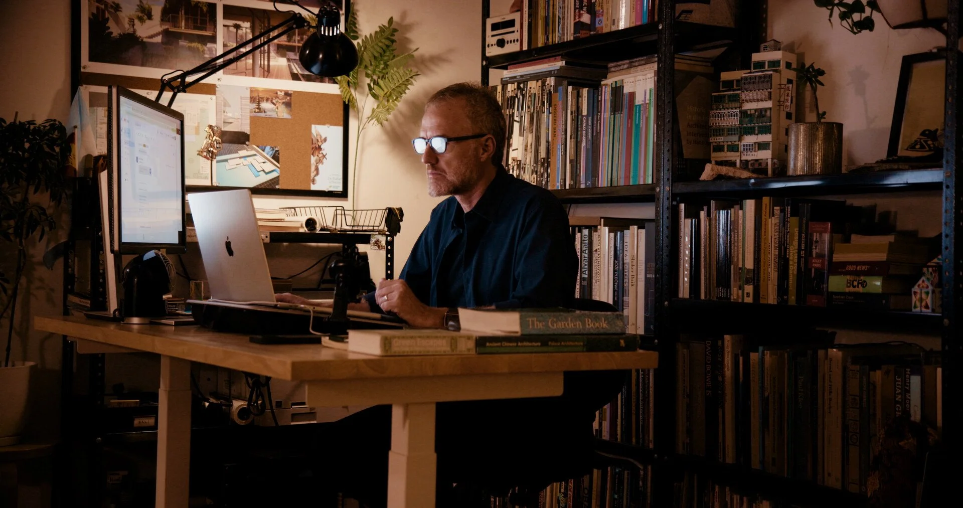 A man wearing glasses working at a desk in a dimly lit room, surrounded by bookshelves filled with books, with a computer monitor and a laptop on the desk.