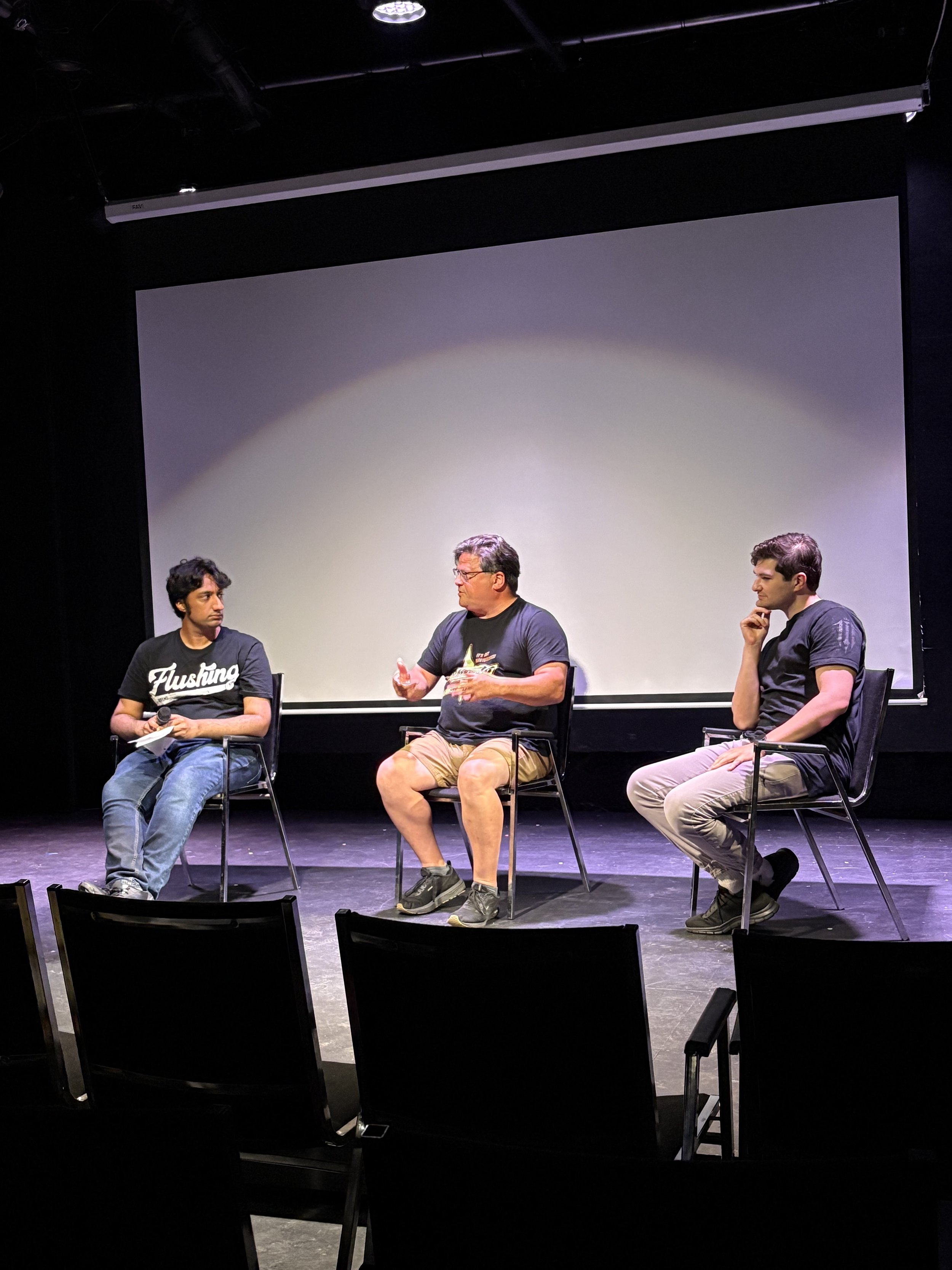Three men sitting on stage in front of a large blank screen, engaged in a discussion. The man in the middle is speaking, holding a bottle, while the other two listen attentively.
