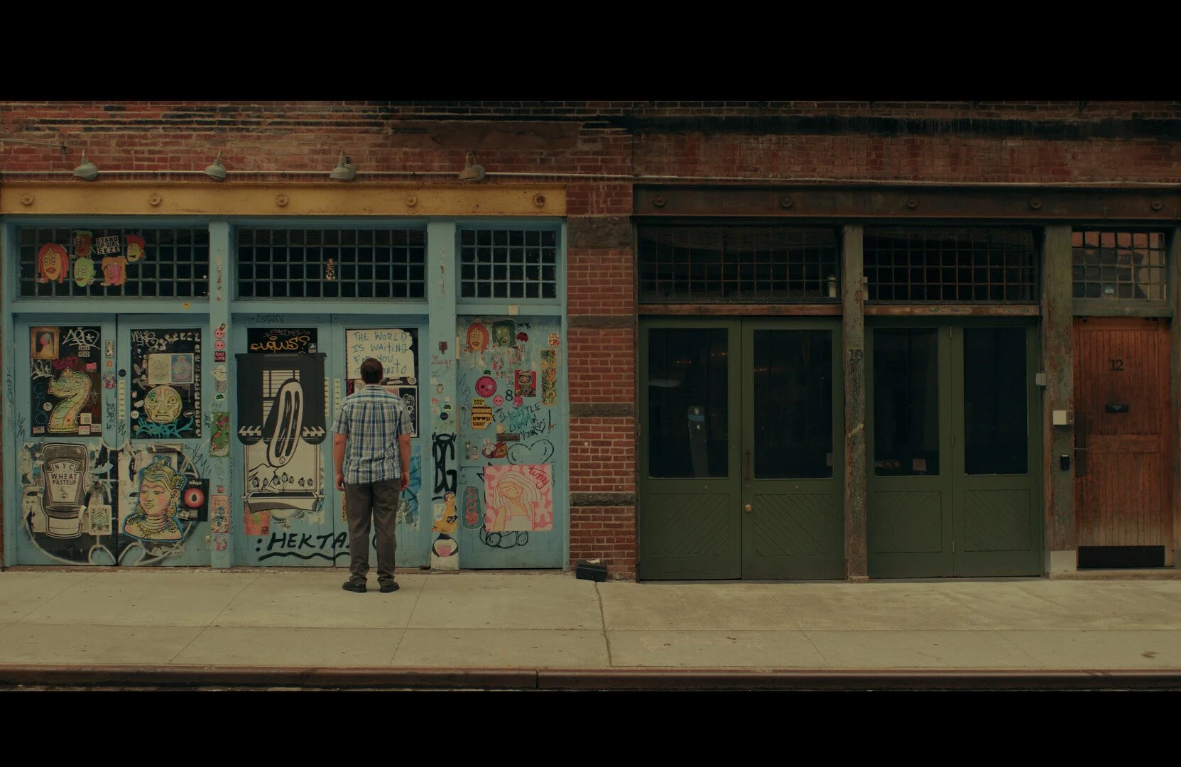 A man in a plaid shirt standing outside a building with a graffiti-covered blue door on the left and a large green door on the right. The wall on the left has various stickers, posters, and artwork.