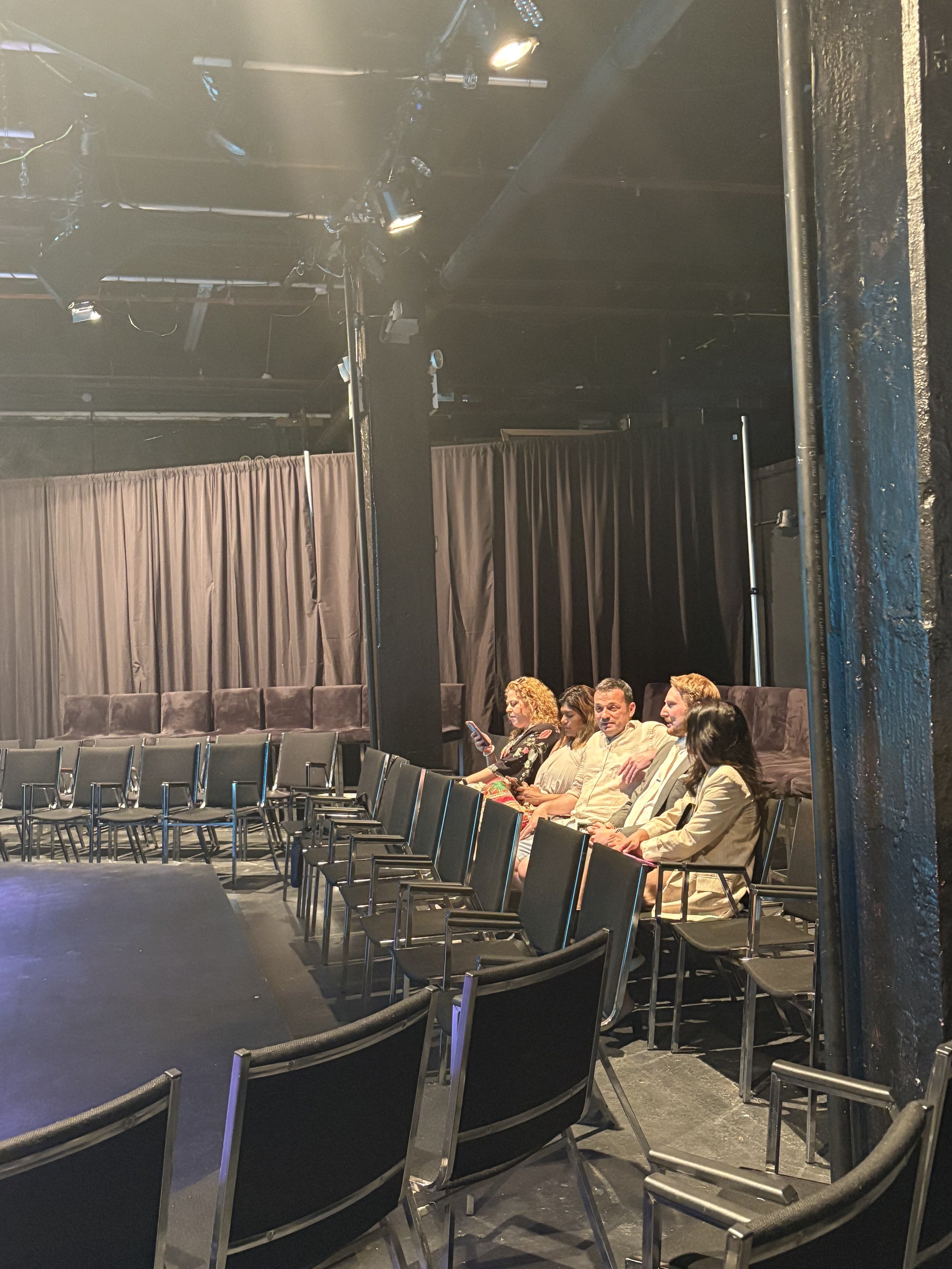 A small audience seated in black chairs in a black-box theater with black curtains and stage lighting.
