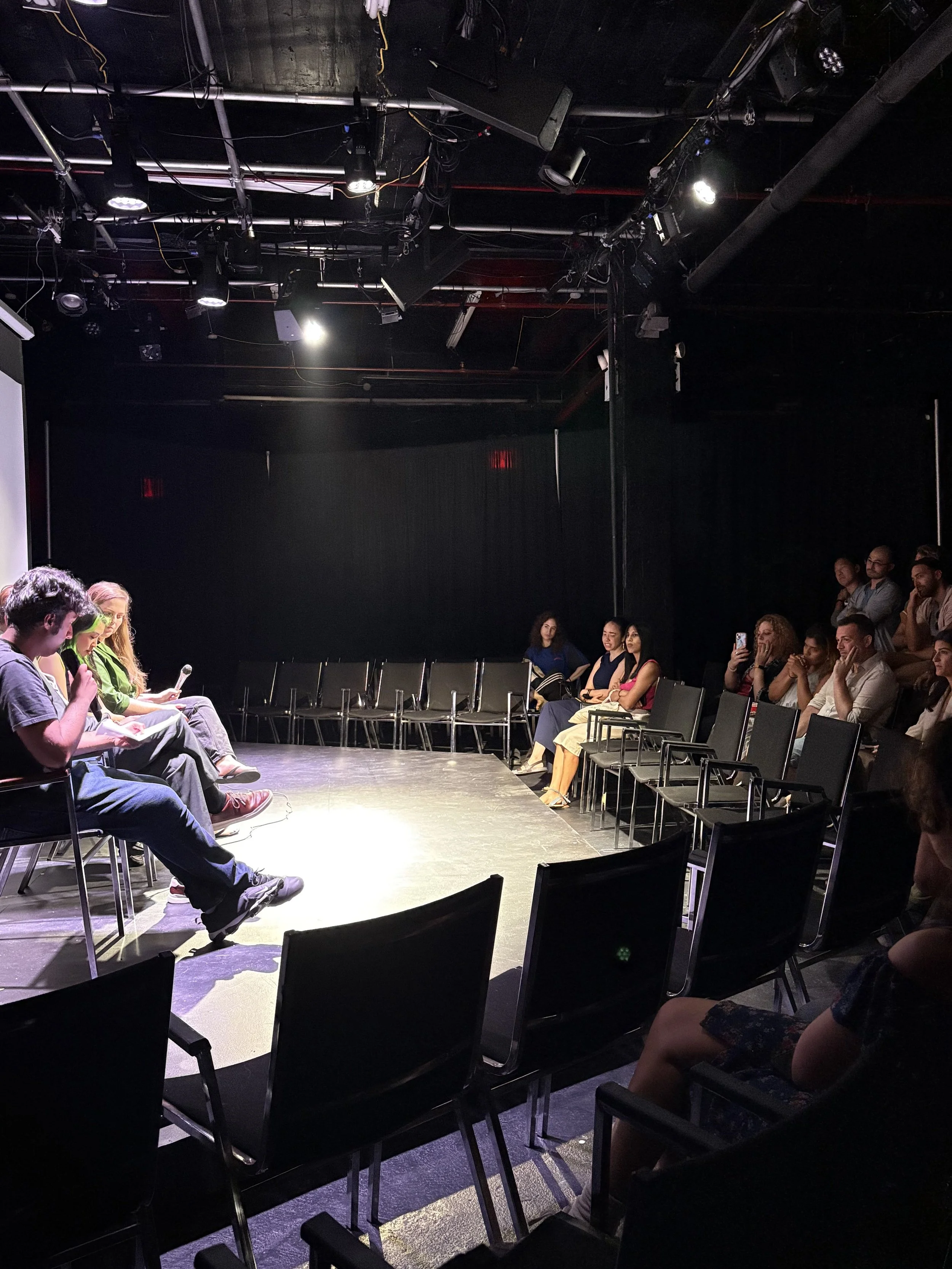 People seated on chairs around a small stage in a black box theater with black walls and ceiling, stage lights overhead, and some audience members looking at scripts or phones.