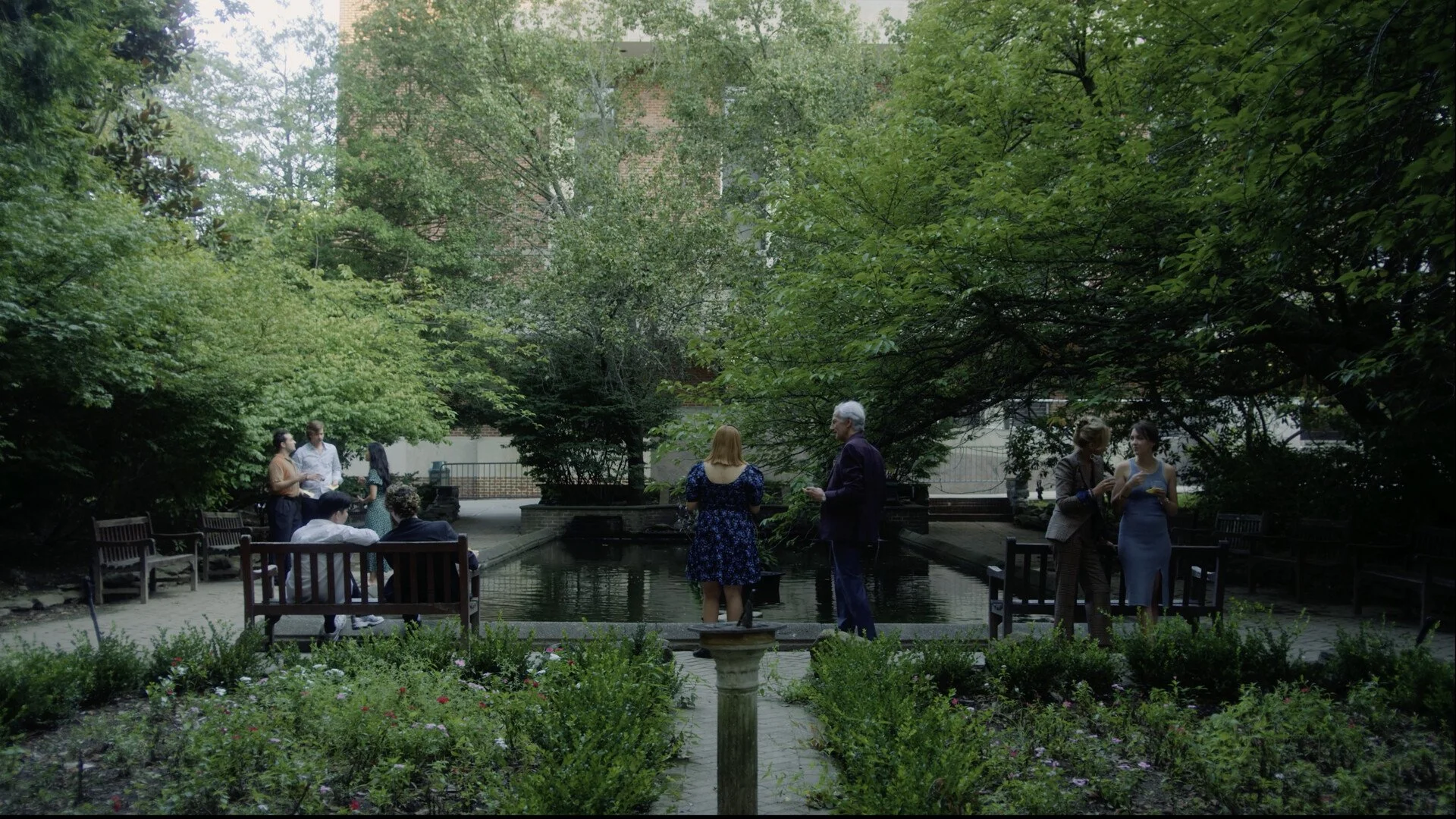 People gathered in a lush green garden with benches and a small fountain, engaging in conversation.