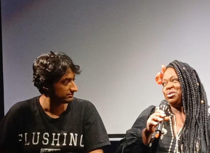 Two people on stage, one holding a microphone and speaking, the other listening attentively, with neutral background.