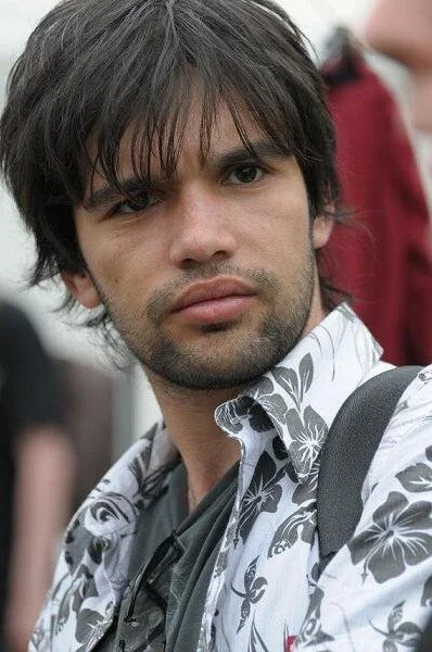 A young man with dark hair and a serious expression, wearing a white shirt with a floral pattern and a backpack, standing outdoors.