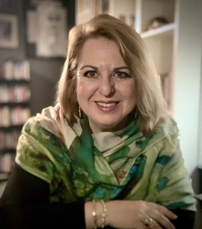 A woman with shoulder-length blonde hair, wearing a colorful scarf, smiling at the camera in a cozy indoor setting with bookshelves in the background.