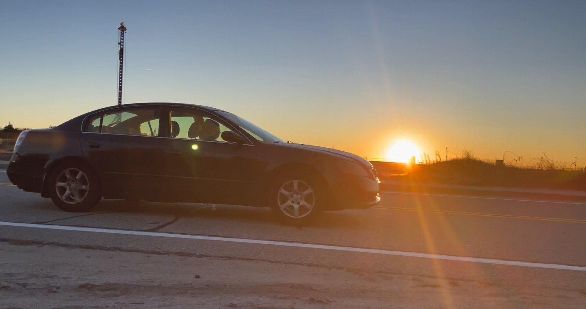 A black sedan parked on the side of a road at sunset, with a clear sky and the sun low on the horizon.