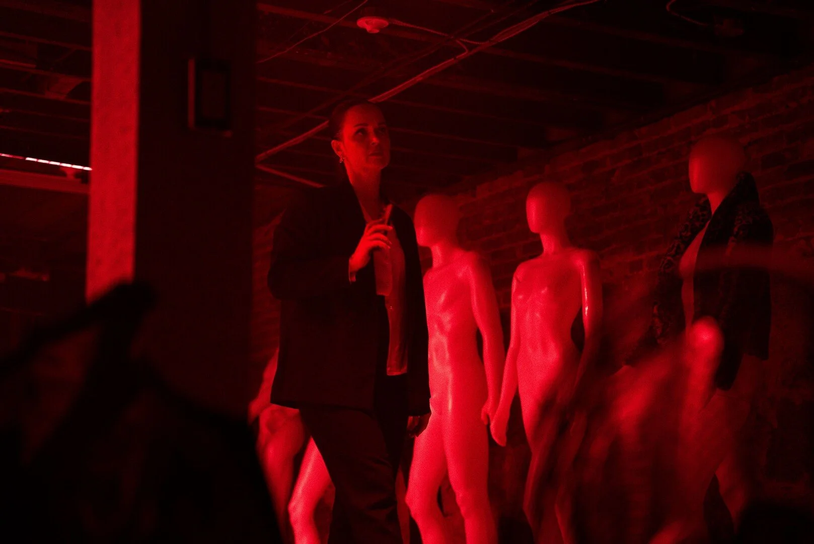 A woman standing in a dark room with red lighting, surrounded by mannequin torsos.
