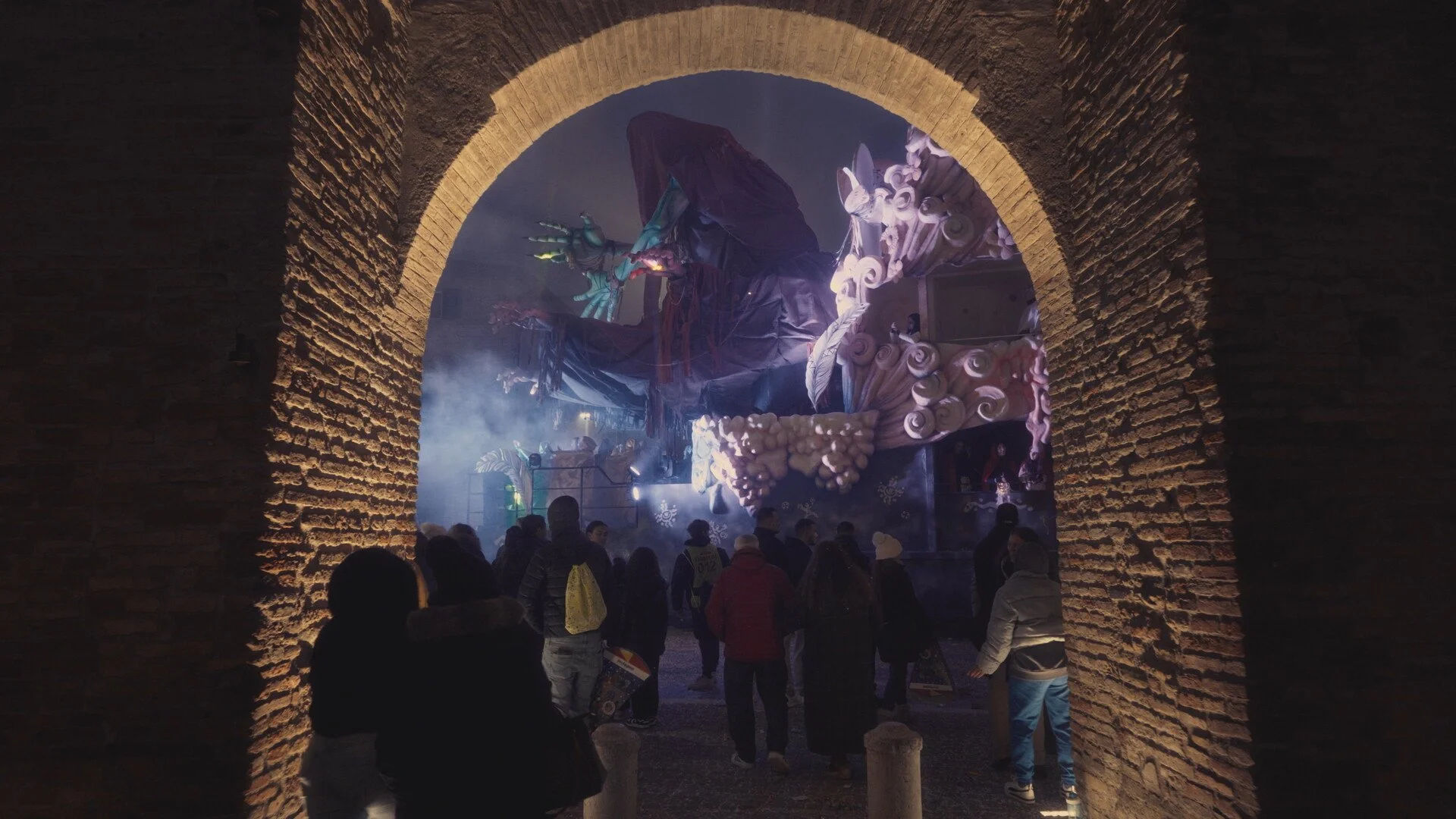 People watching a large, illuminated, dragon-themed parade float through an arched brick passageway at night.