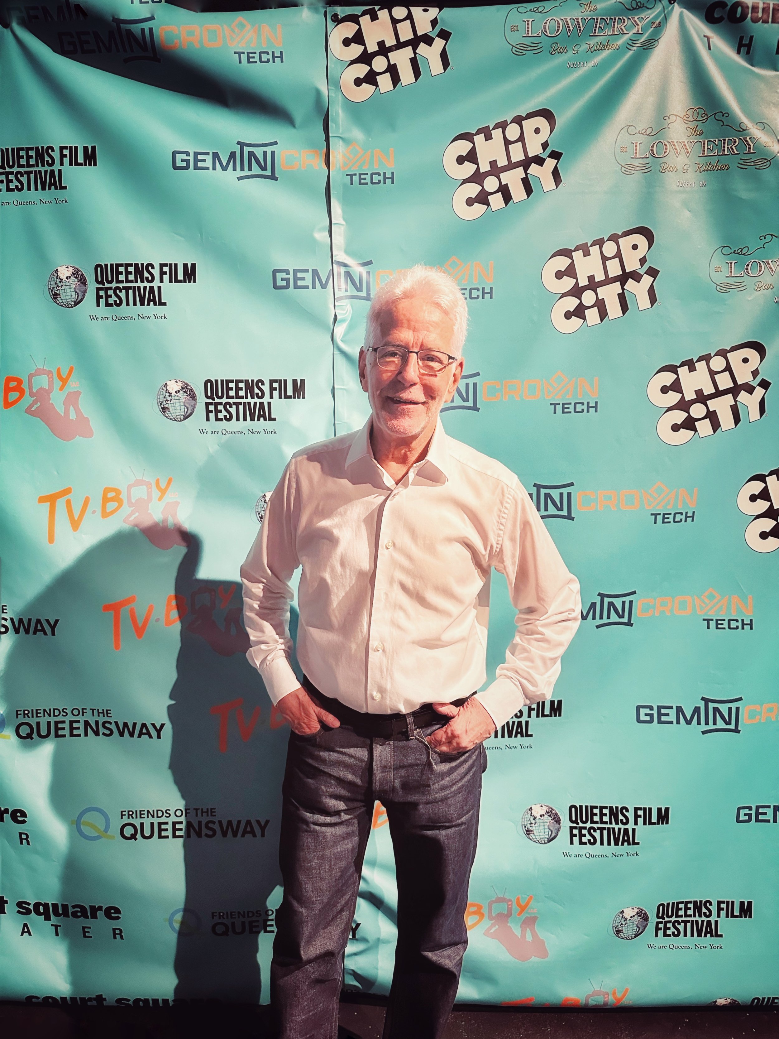 An elderly man with white hair, glasses, wearing a white shirt and gray pants, standing with his hands in his pockets, smiling at the camera in front of a teal backdrop with various event logos and text.