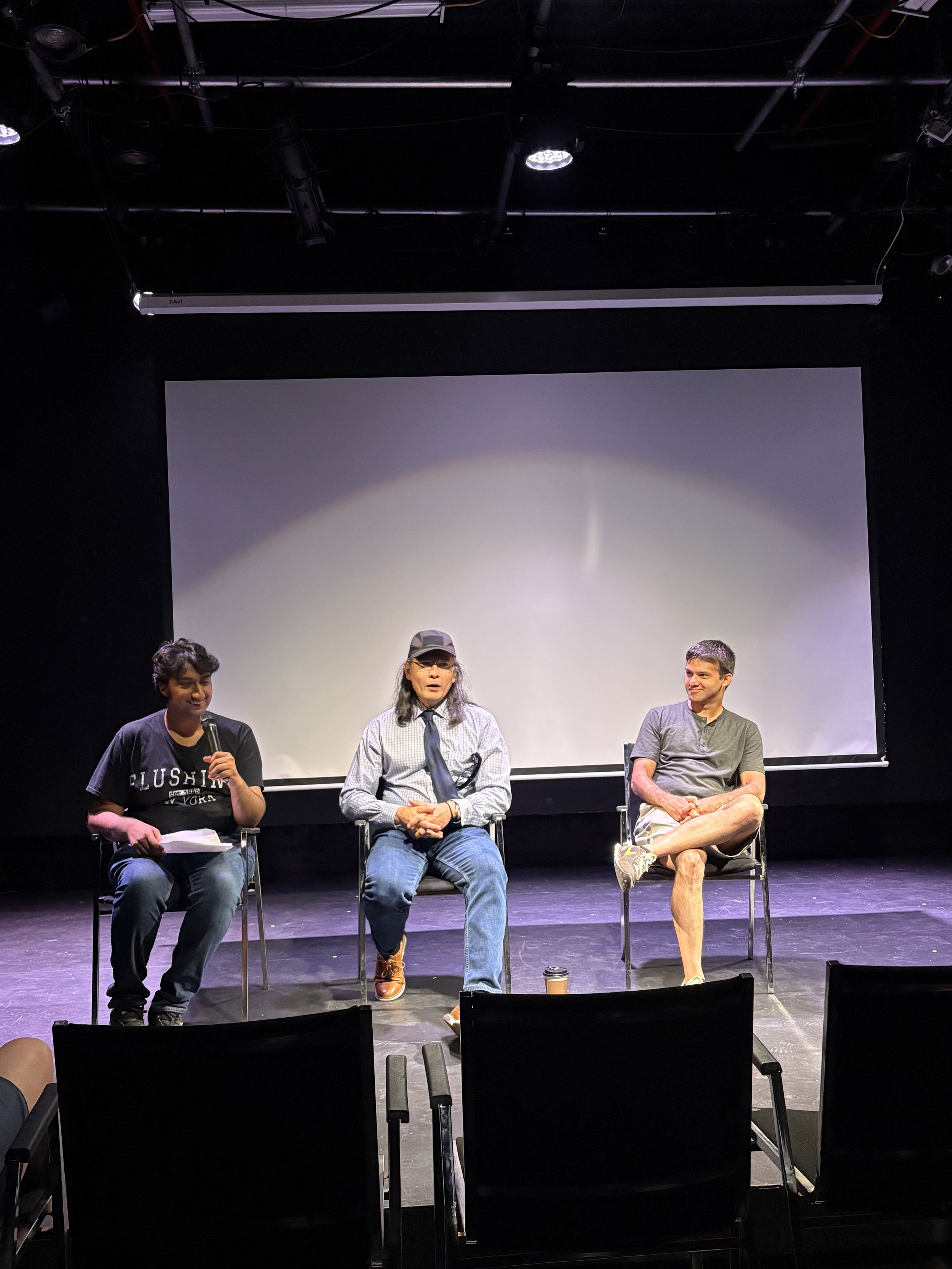 Three people sit on chairs on a stage with a large blank screen behind them. The person on the left has short curly hair and is speaking into a microphone. The person in the middle has long hair, glasses, and wears a cap. The person on the right has short hair and is smiling with legs crossed.