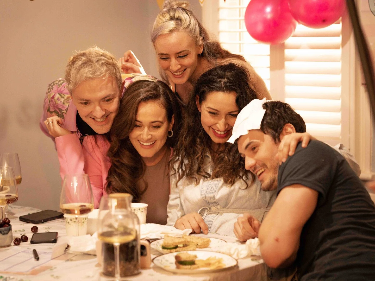 Group of friends gathered around a table, smiling and looking at a man with a head injury covered with a tissue, during a casual indoor celebration or get-together.