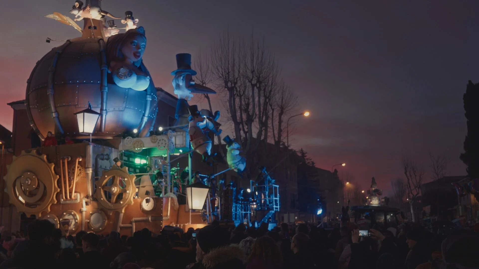 Nighttime parade with large, colorful floats featuring characters, including a woman with a large head and other animated figures, surrounded by a crowd of spectators.