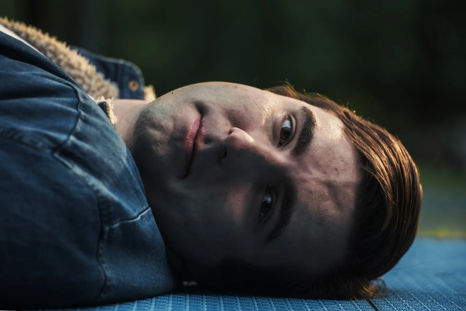 Close-up of a young man lying on his side on a blue surface outdoors with a dark background, looking at the camera with a calm expression.